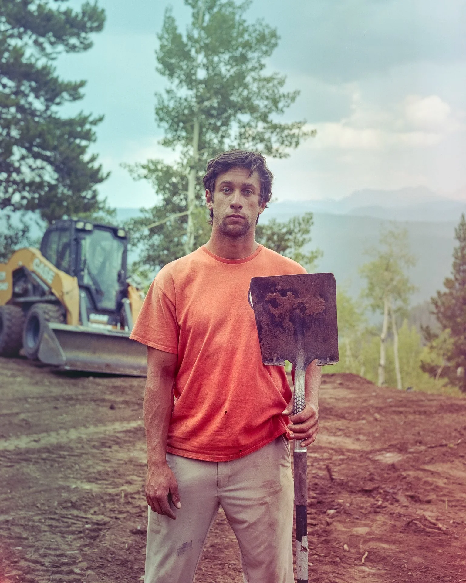 A man with dark hair, wearing an orange T-shirt and beige pants, standing outdoors holding a shovel stained with dirt. There is a backhoe loader in the background, and the setting appears to be a construction or landscaping site in a natural, wooded 