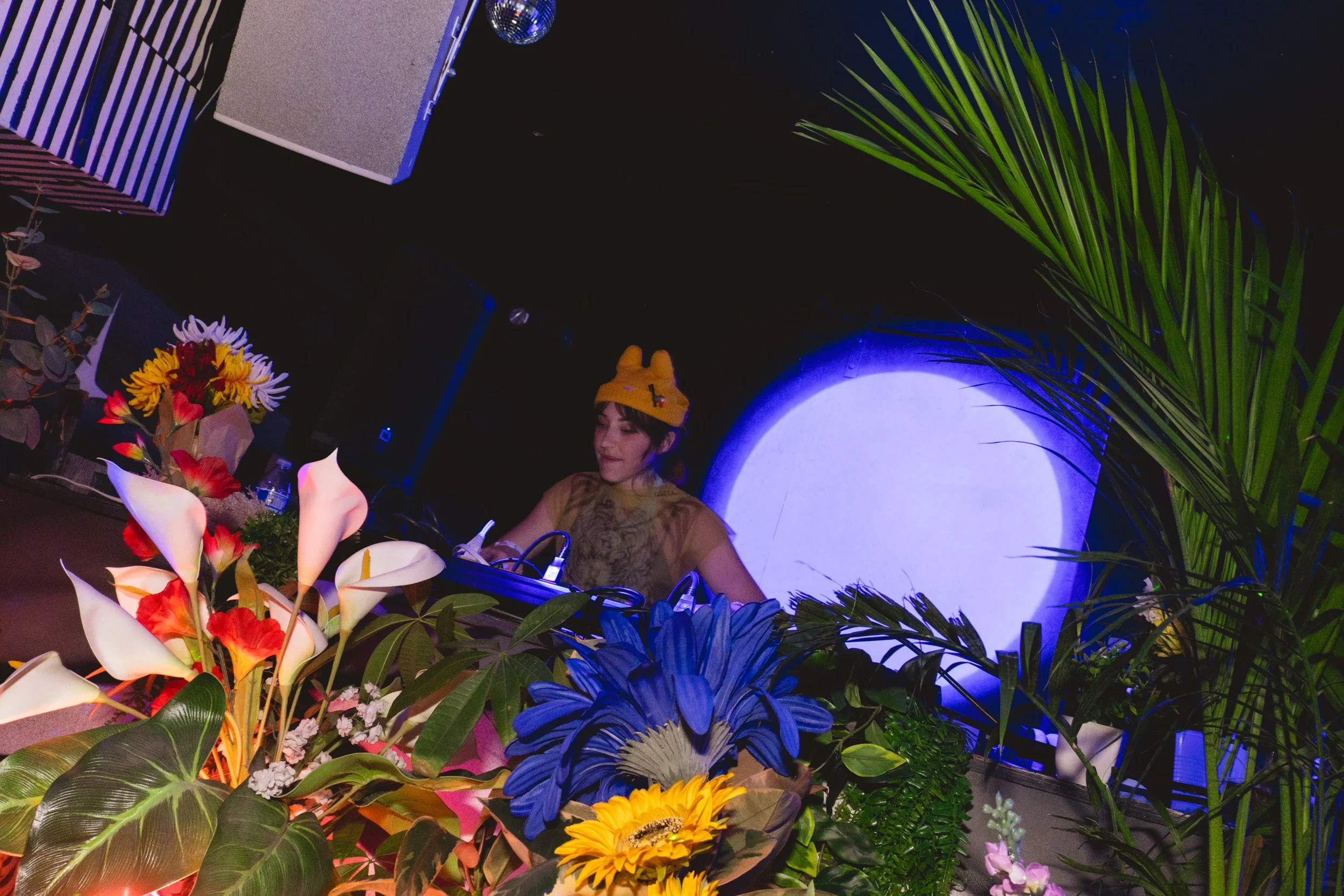 A woman DJ wearing a yellow hat with a bunny design is performing at a venue decorated with colorful flowers and green plants, with a large blue and white circular light in the background.