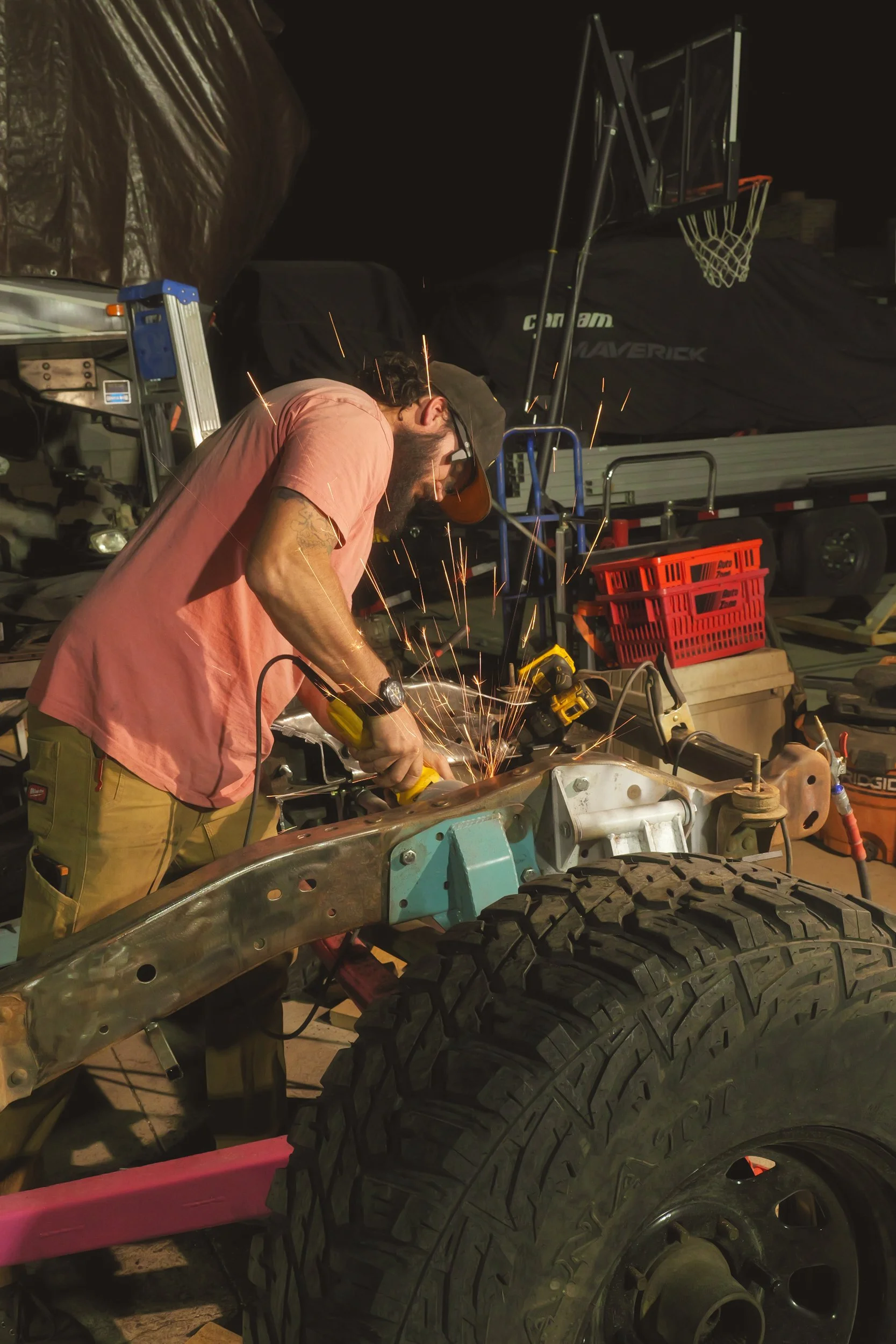 A man welding a metal frame with sparks flying in a workshop filled with tools and equipment, including a large tire in the foreground.