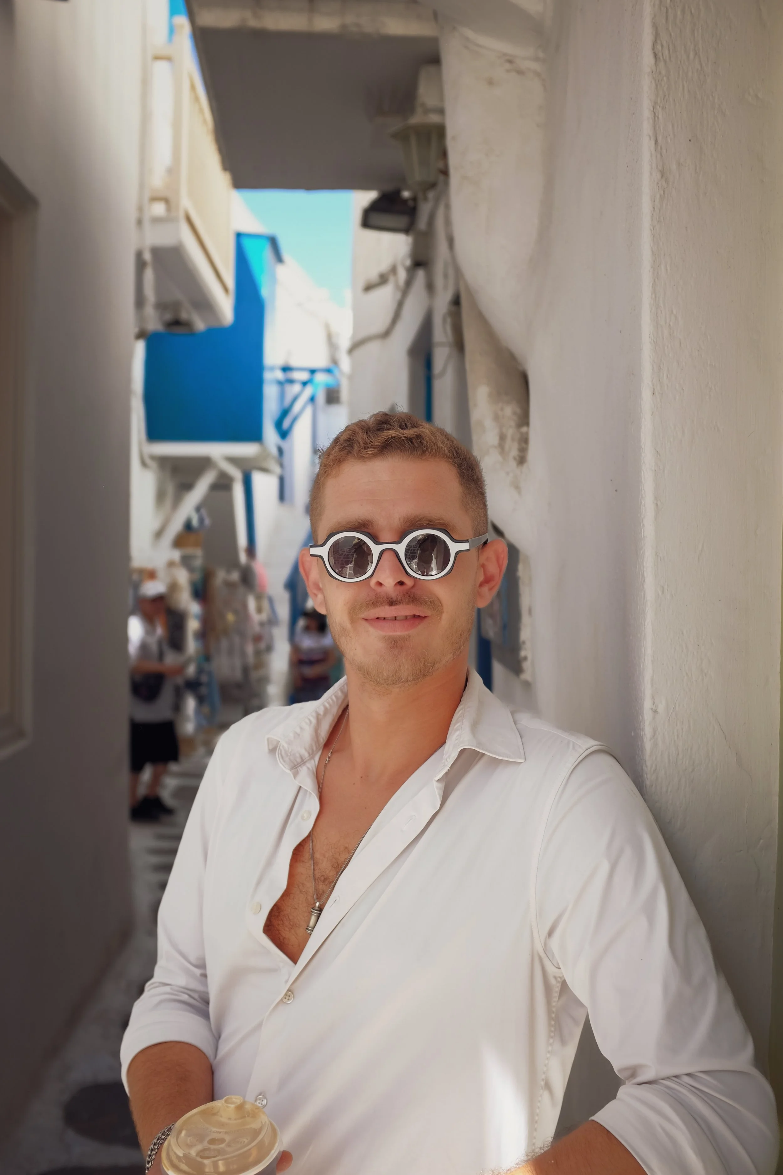 A young man with light skin, short curly hair, wearing round sunglasses, a white open-collared shirt, holding a coffee cup, standing in a sunlit alley with white and blue buildings and blurred people in the background.