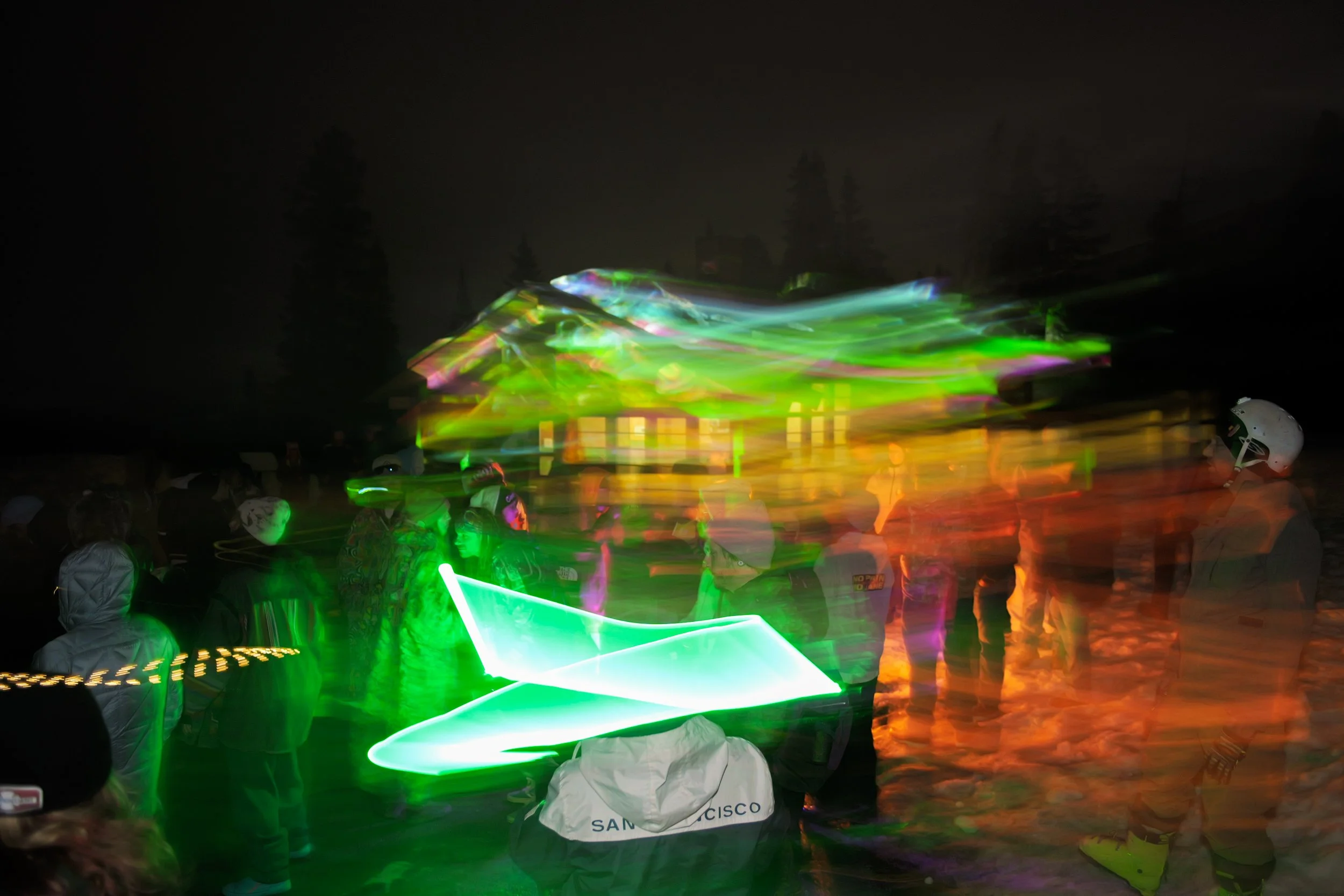 Nighttime outdoor scene with people, some wearing helmets, gathered around illuminated green and white light shapes, with a house and trees faintly visible in the background.