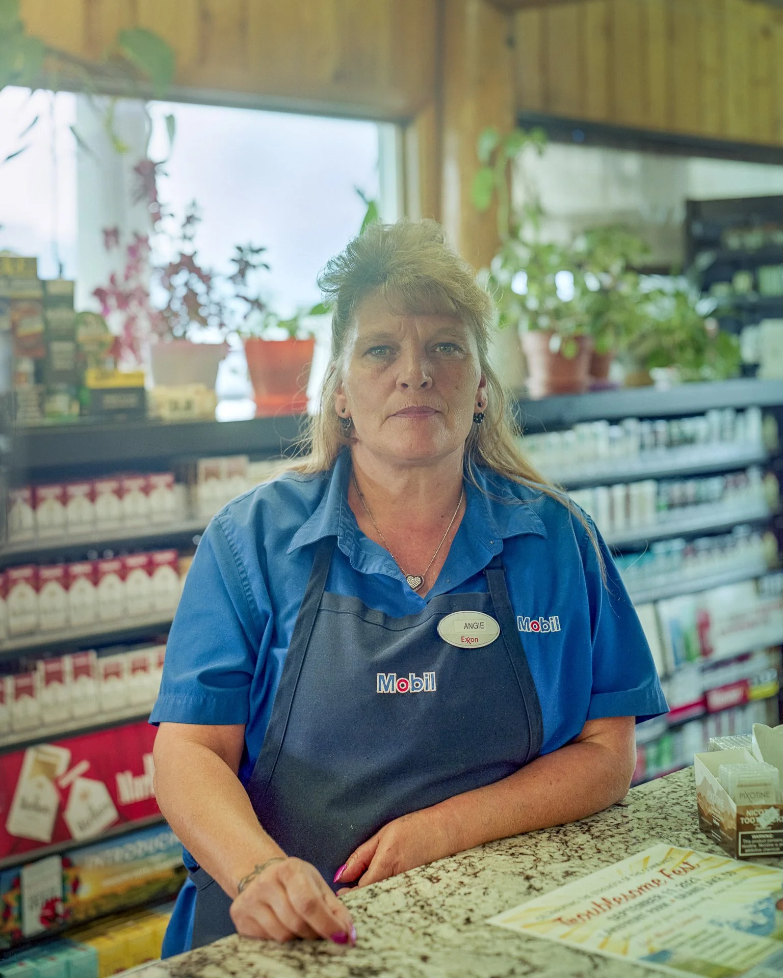 A woman with blonde hair and a serious expression, wearing a blue shirt and a Mobil apron, standing behind a counter in a convenience store with shelves of products and plants in the background.
