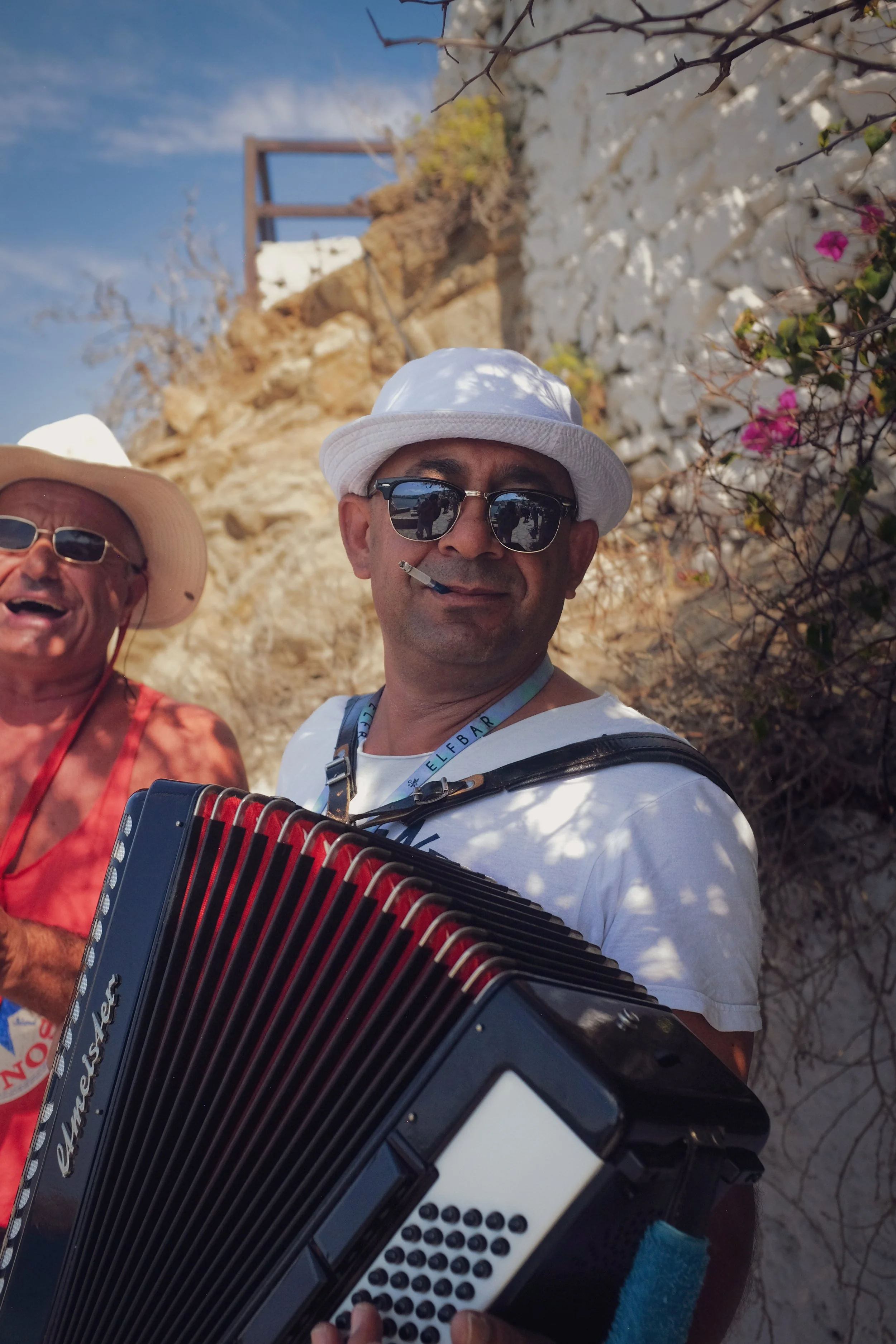 Two men wearing white hats and sunglasses, one playing an accordion, standing outdoors against a rocky background and flowering plants with pink flowers.