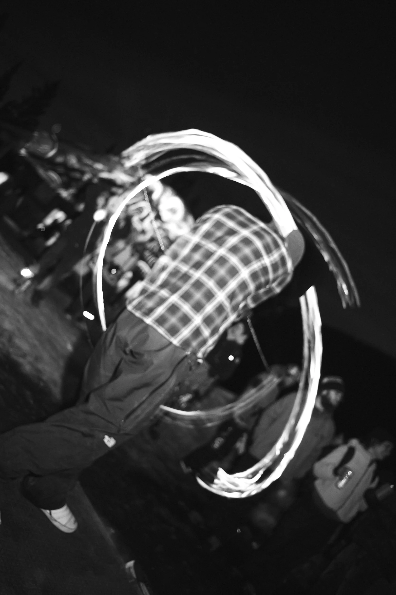 A man is spinning a light trail in a dark outdoor setting, creating a circular light effect, with people in the background.