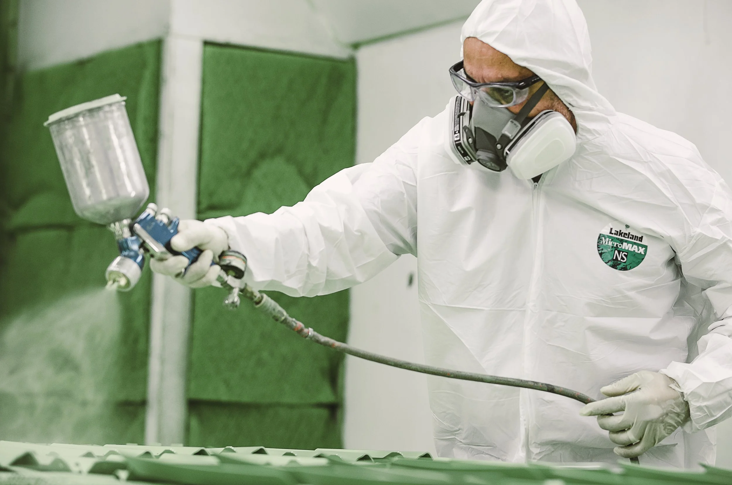 Person wearing protective gear and mask spray painting or coating a surface in an industrial or manufacturing setting.