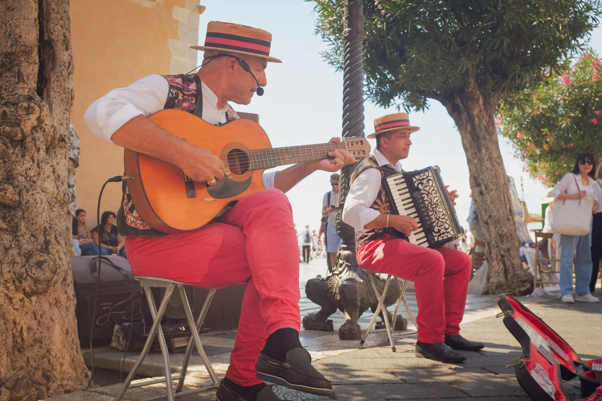Two street musicians wearing traditional hats and red pants, one playing guitar and the other accordion, perform outdoors under trees with spectators nearby.