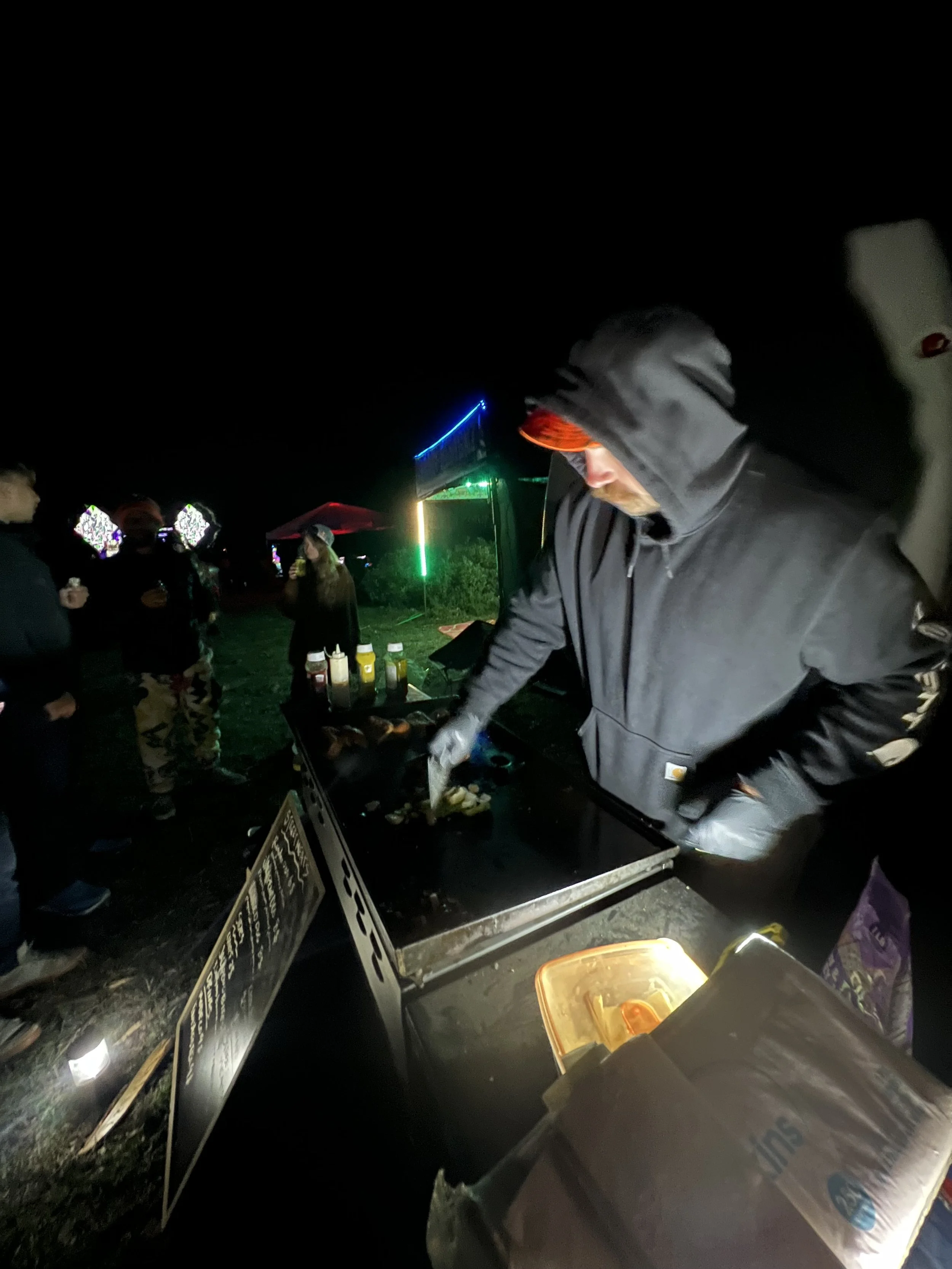 Person in a black hoodie cooking food at an outdoor night event with a group of people and colorful lights in the background.