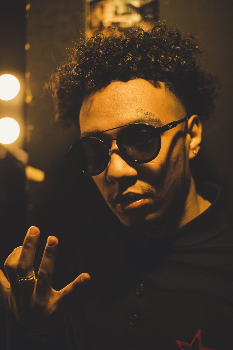 A person with curly hair, wearing sunglasses and a dark jacket, making a peace sign gesture.