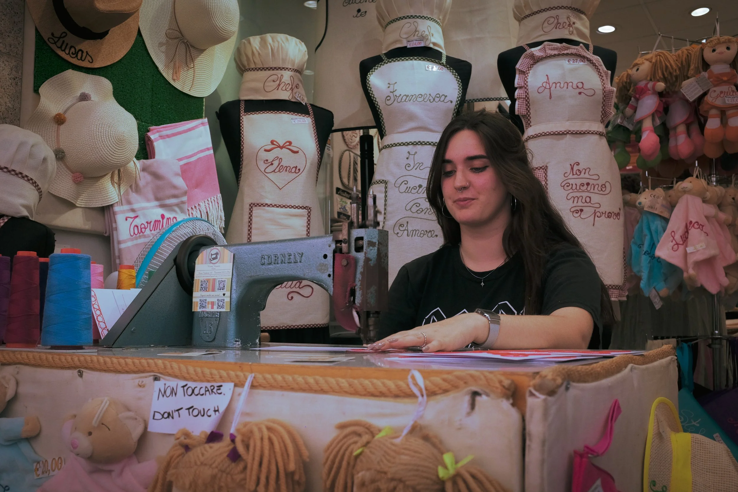 Young woman working at a sewing or embroidery station with various colorful spools of thread, plush toys, and embroidered aprons with Italian phrases in the background.