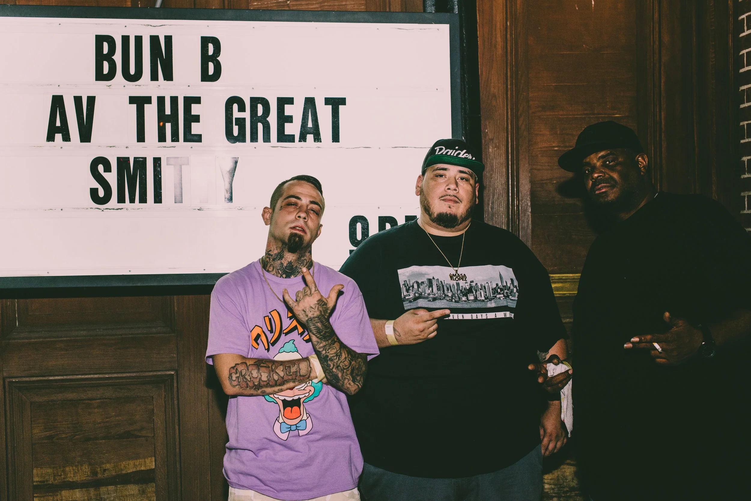 Three men standing in front of a white sign with black text. The sign says "BUN B AV THE GREAT SMIT" with some letters obscured. The men are posing for the camera, one wearing a purple shirt with cartoon character print, tattoos on his arms, and maki