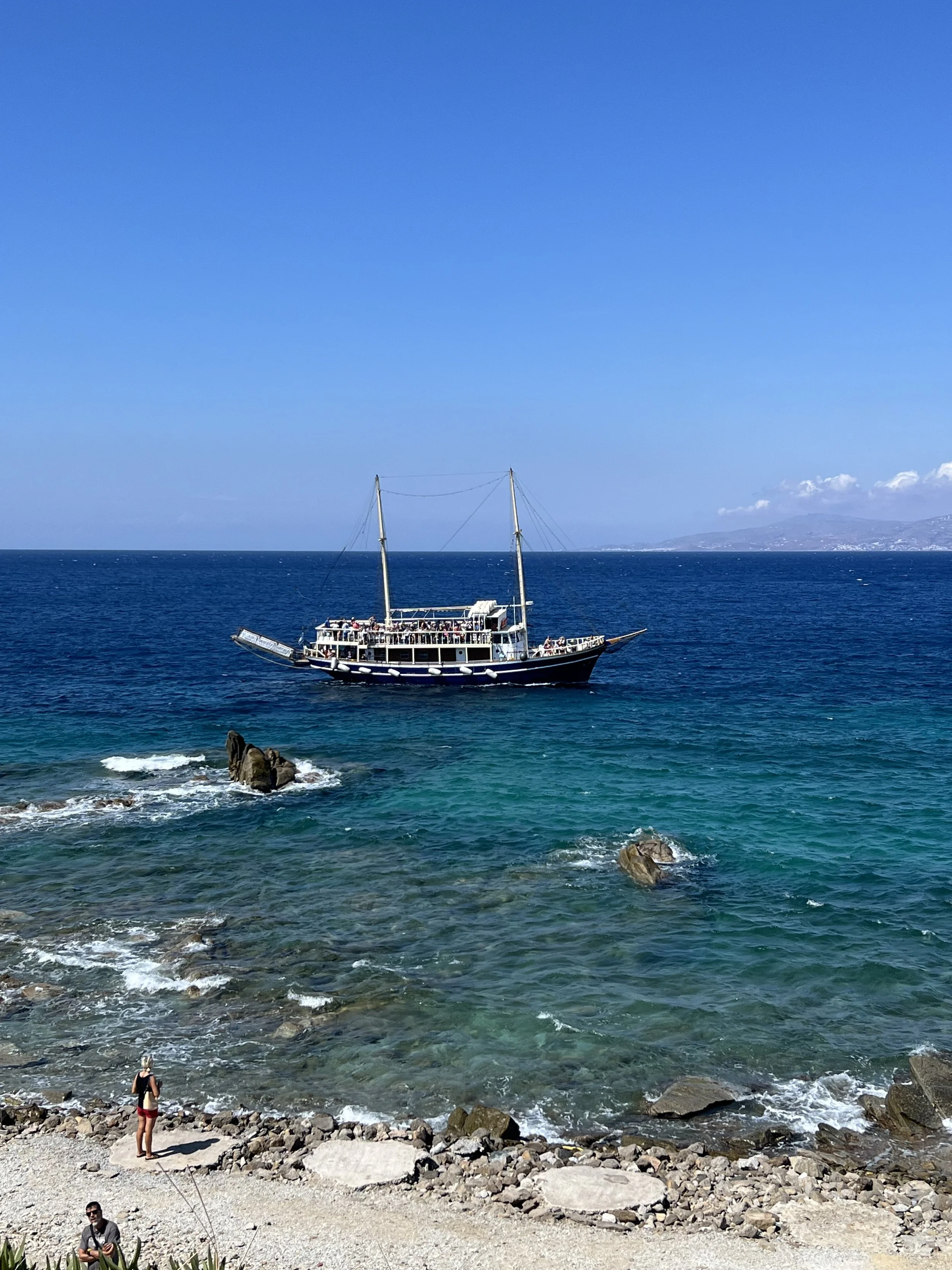 A large sailing ship with multiple decks and two masts sailing on the ocean near a rocky shoreline. There are a few people on the beach, some taking photos, and a person in the foreground holding a fishing rod. The sky is clear with a few clouds in t