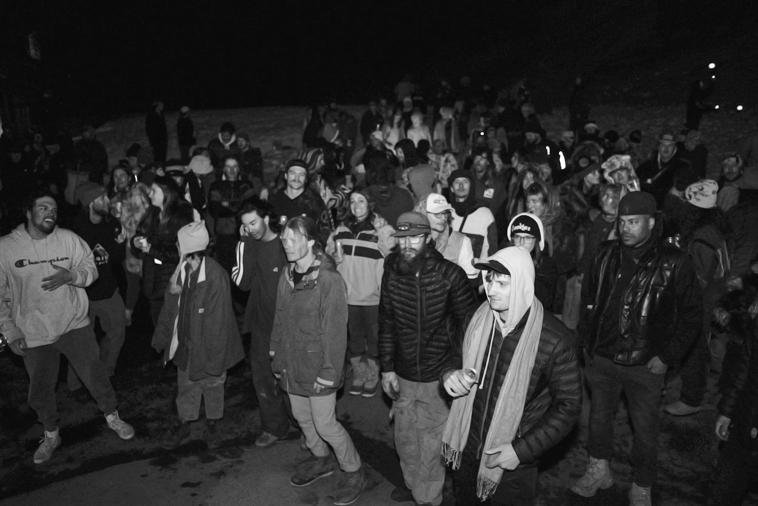 A large group of people gathered outdoors at night, some holding drinks, standing and socializing.
