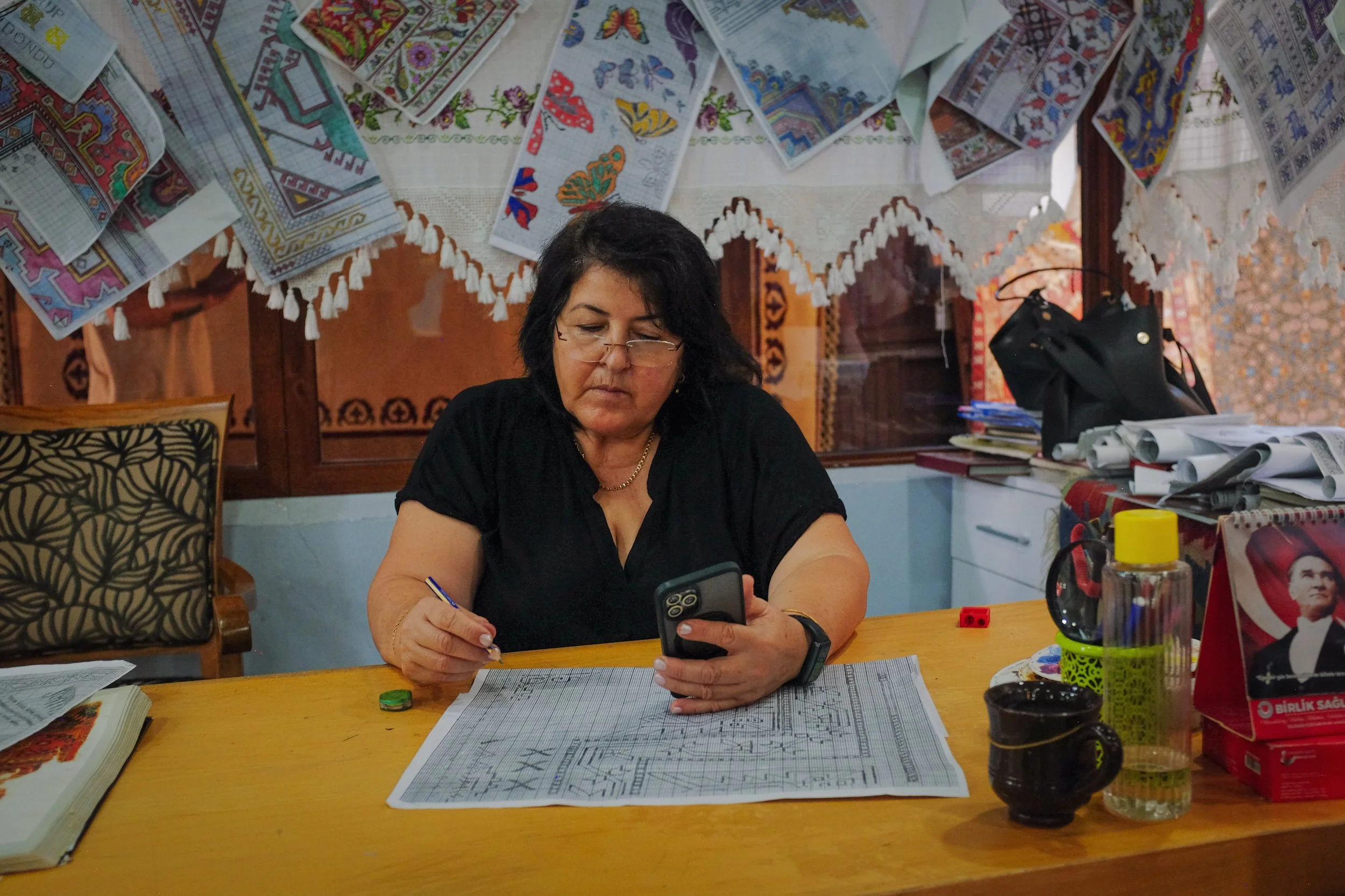A woman with dark hair and glasses sitting at a wooden table, looking at her phone, with architectural plans spread in front of her, surrounded by papers, books, and office supplies, with embroidered and colorful textiles hanging on the wall behind h