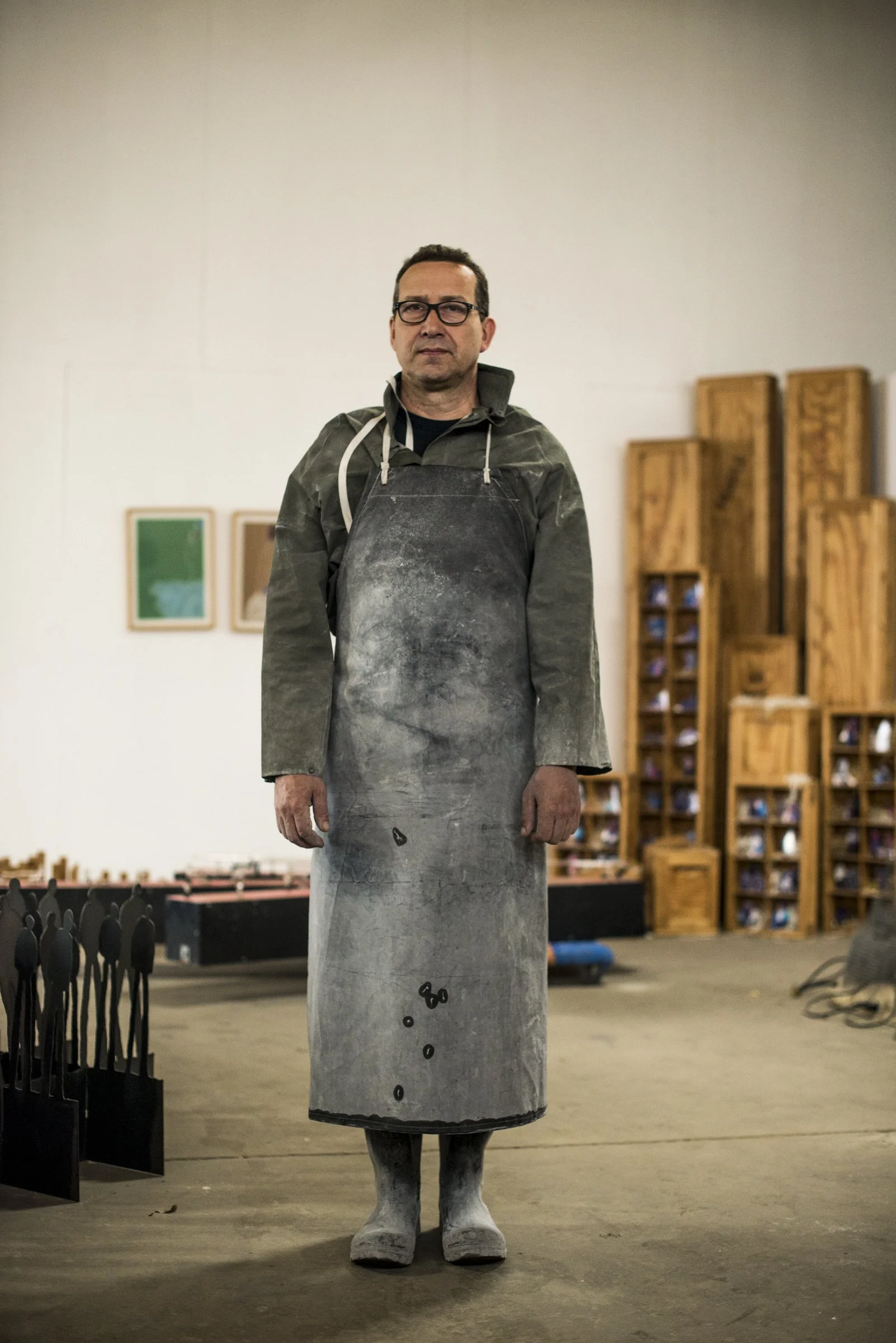 Man standing indoors in a white-room, wearing glasses, a dark jacket, and an apron with paint or chalk stains, with wooden furniture and framed pictures in the background.