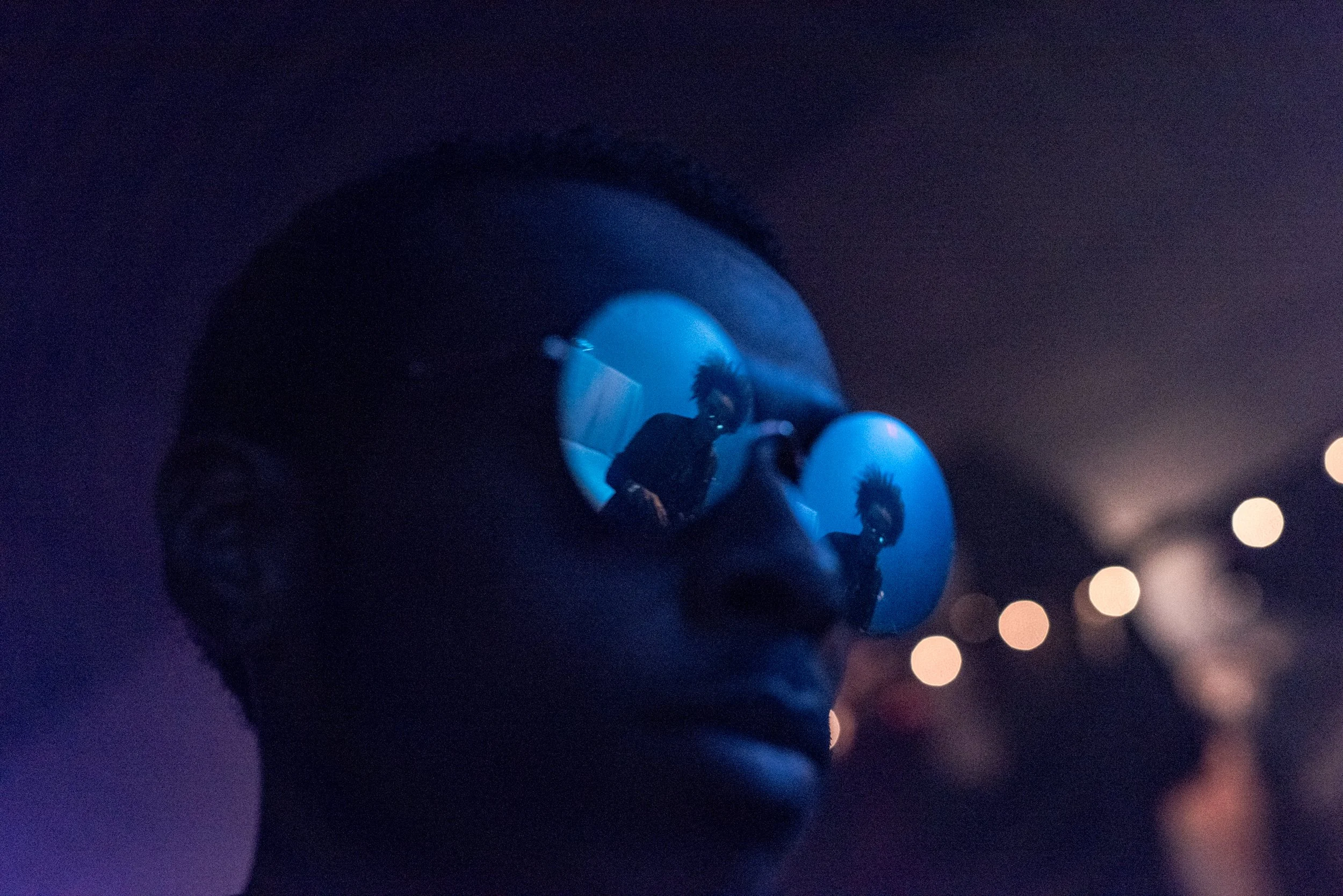 A person wearing reflective sunglasses with a blue tint, reflecting a woman with wild hair and a dark outfit in a dimly lit environment with bokeh lights in the background.