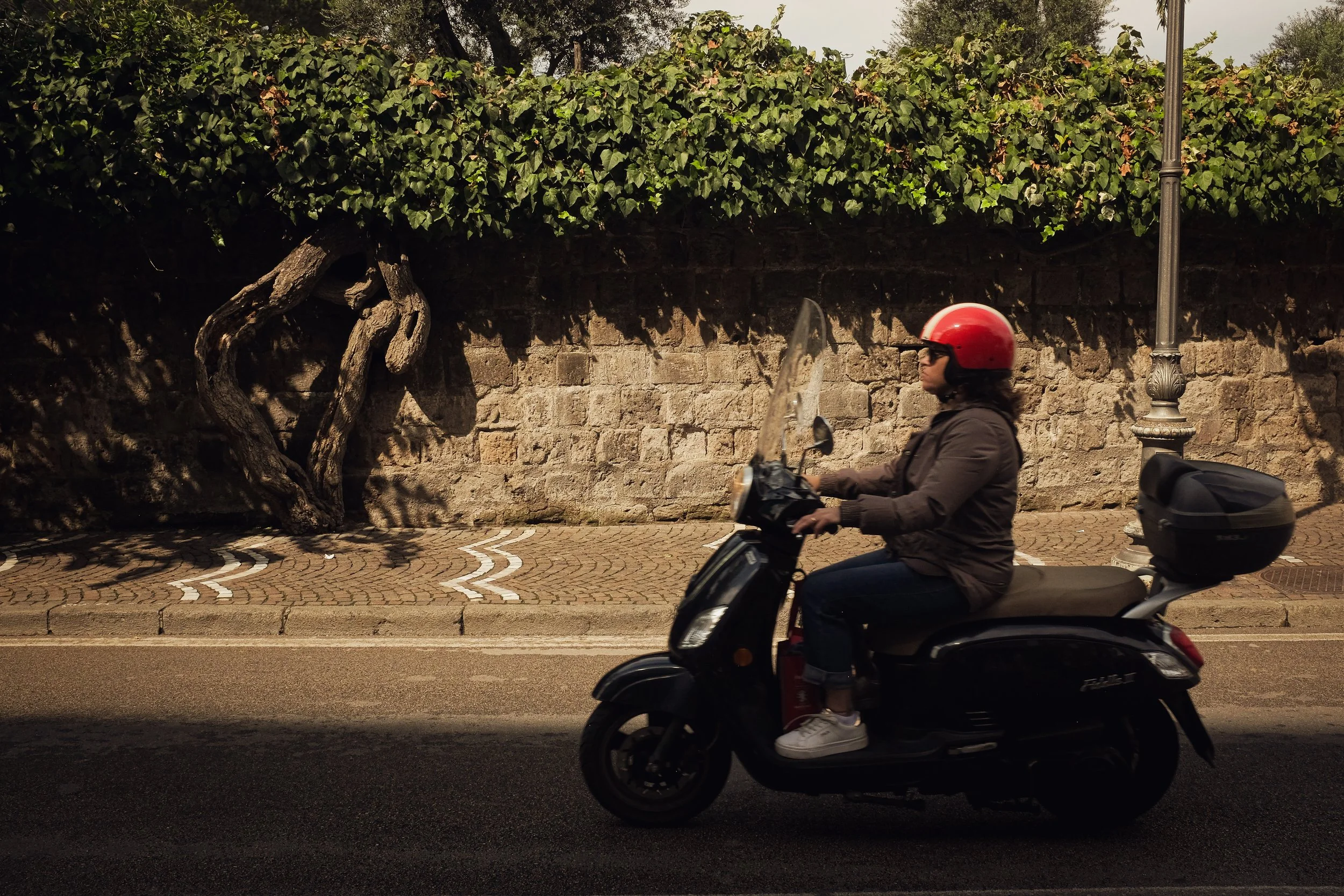 A person riding a black scooter while wearing a red helmet on a paved street next to a stone wall with ivy on top.
