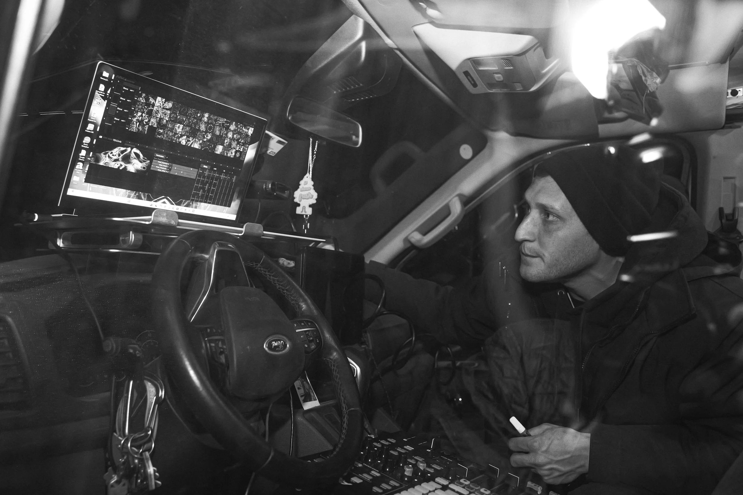 A man is sitting in the driver's seat of a truck. There is a laptop on the dashboard displaying a photo editing or music software. The man is wearing a beanie and a jacket, holding a cigarette, and looking to the right.