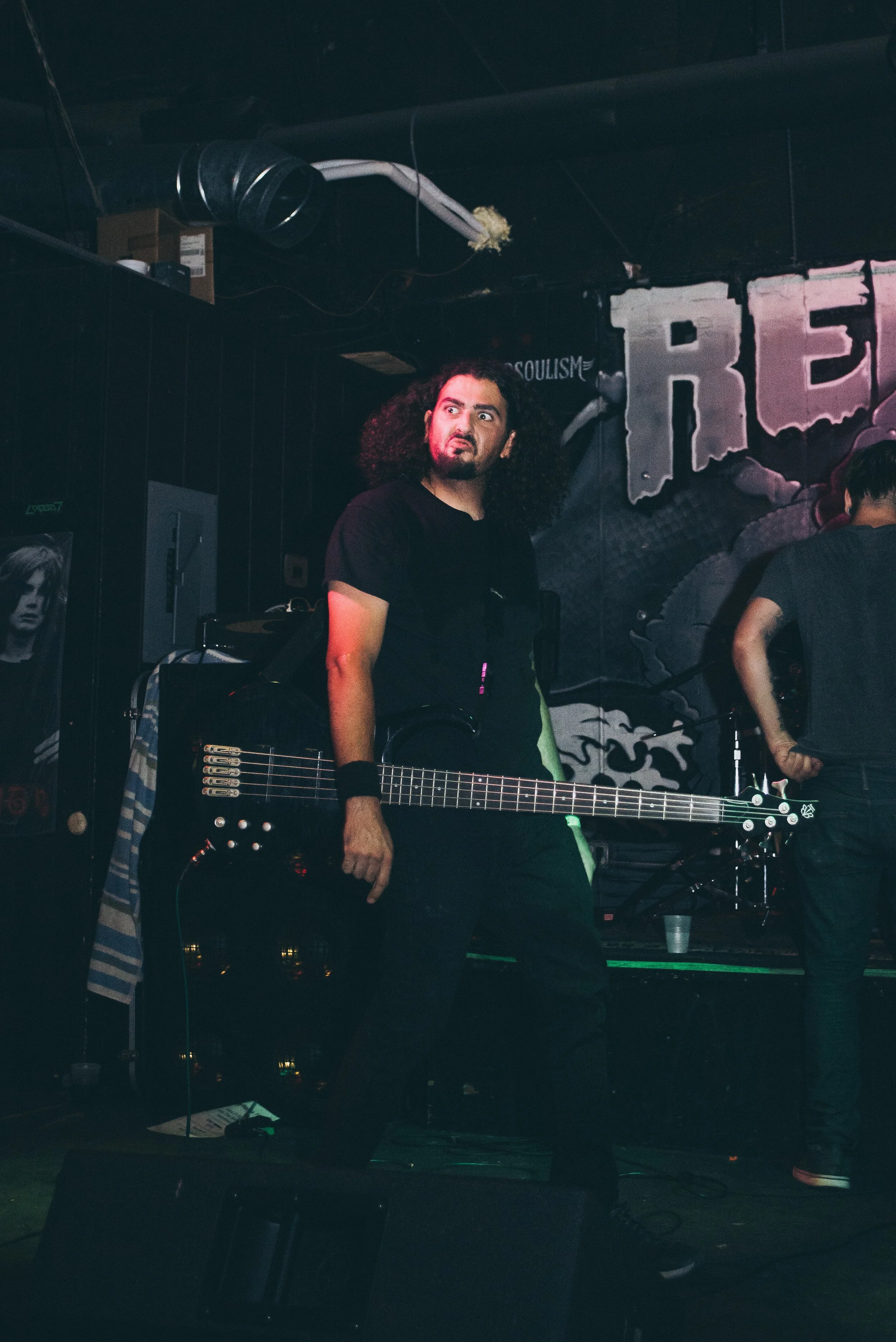 A man with curly hair holding a bass guitar on a stage, with dark background and colorful stage lights.