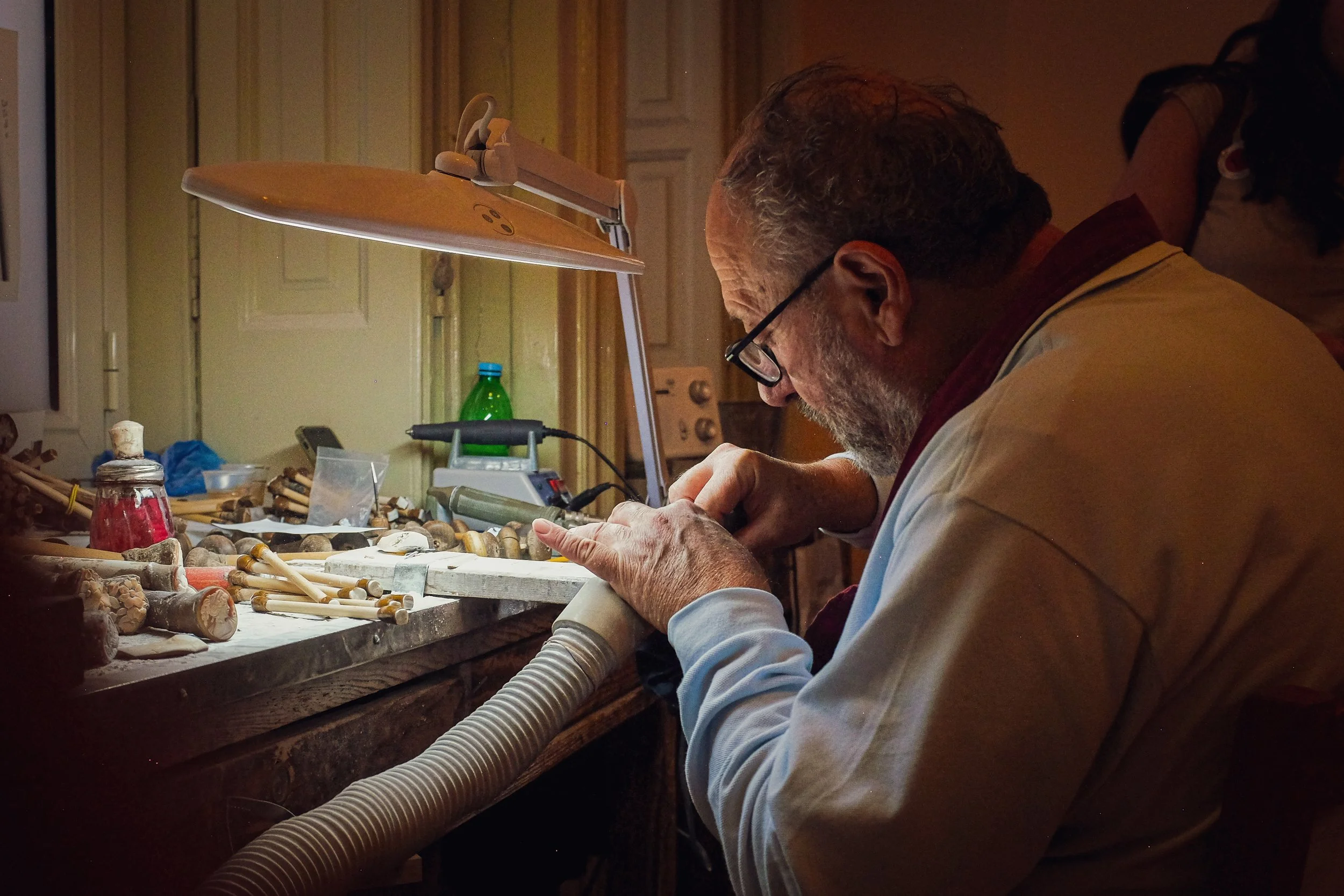 An elderly man with glasses working on a woodworking or carving project at a cluttered workbench with tools and wood pieces, using a vacuum hose.