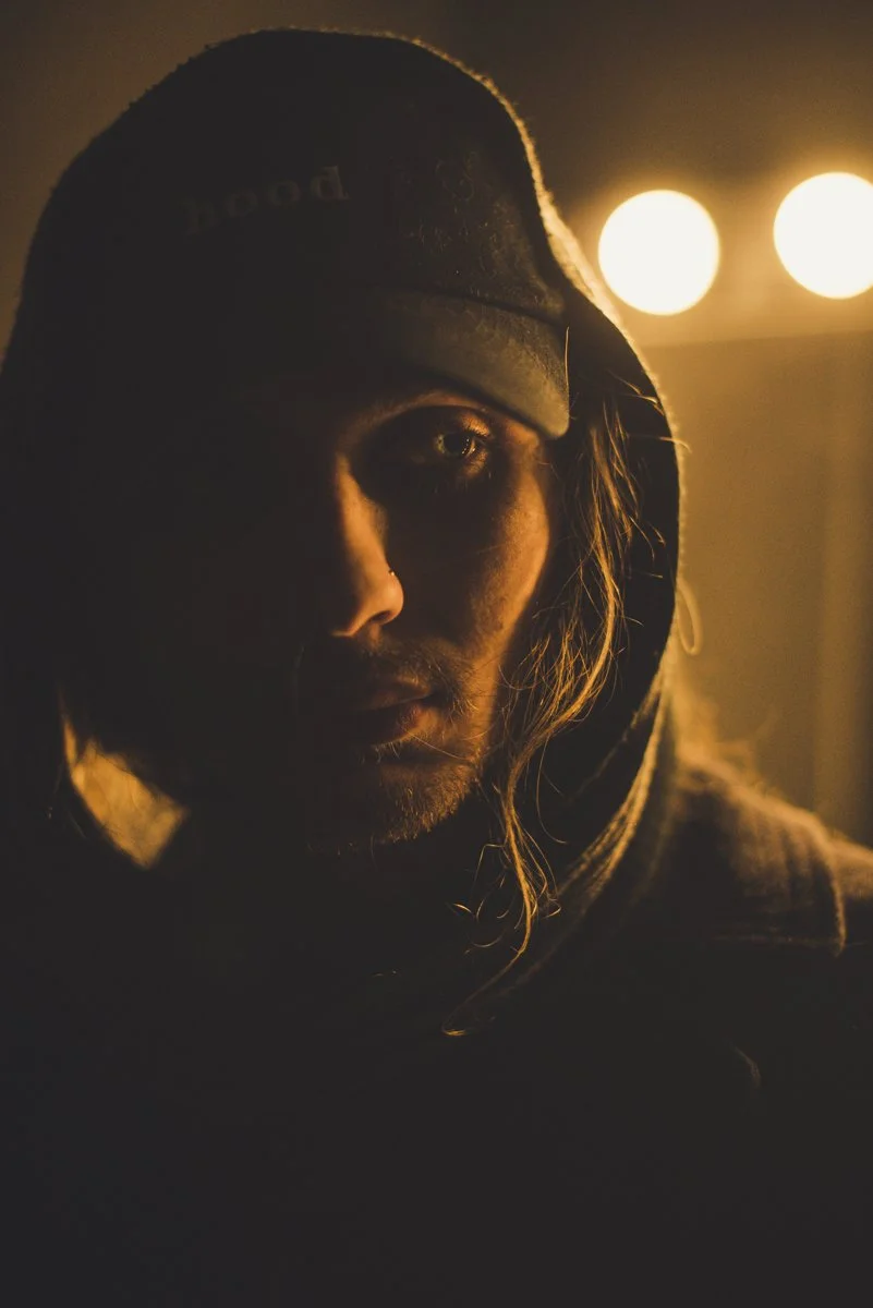 A person with long hair wearing a hood and cap, lit by warm, low lighting in a dark environment.