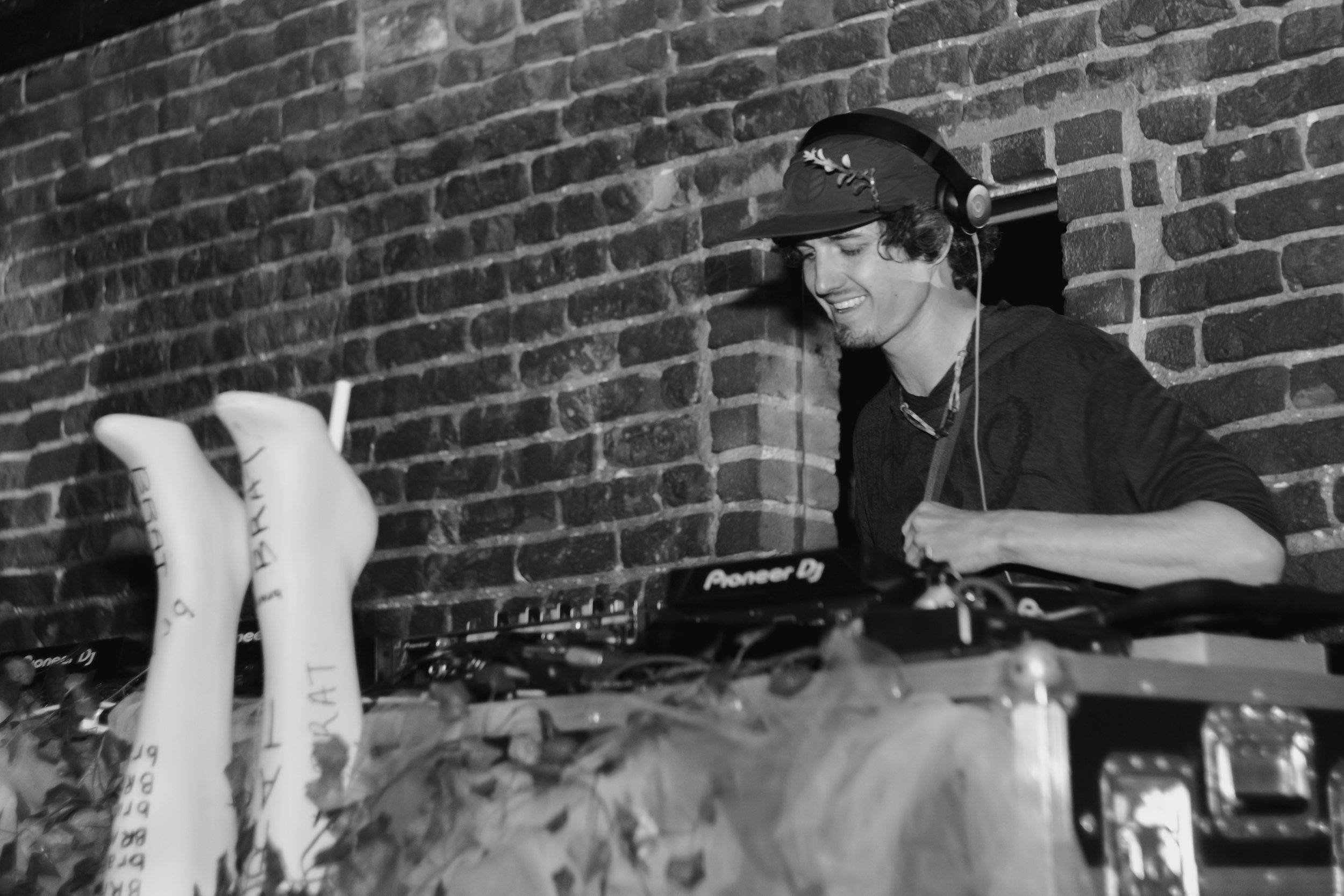 A young man smiling, wearing headphones, a hat, and a dark shirt, working as a DJ with turntables and DJ equipment in front of a brick wall.