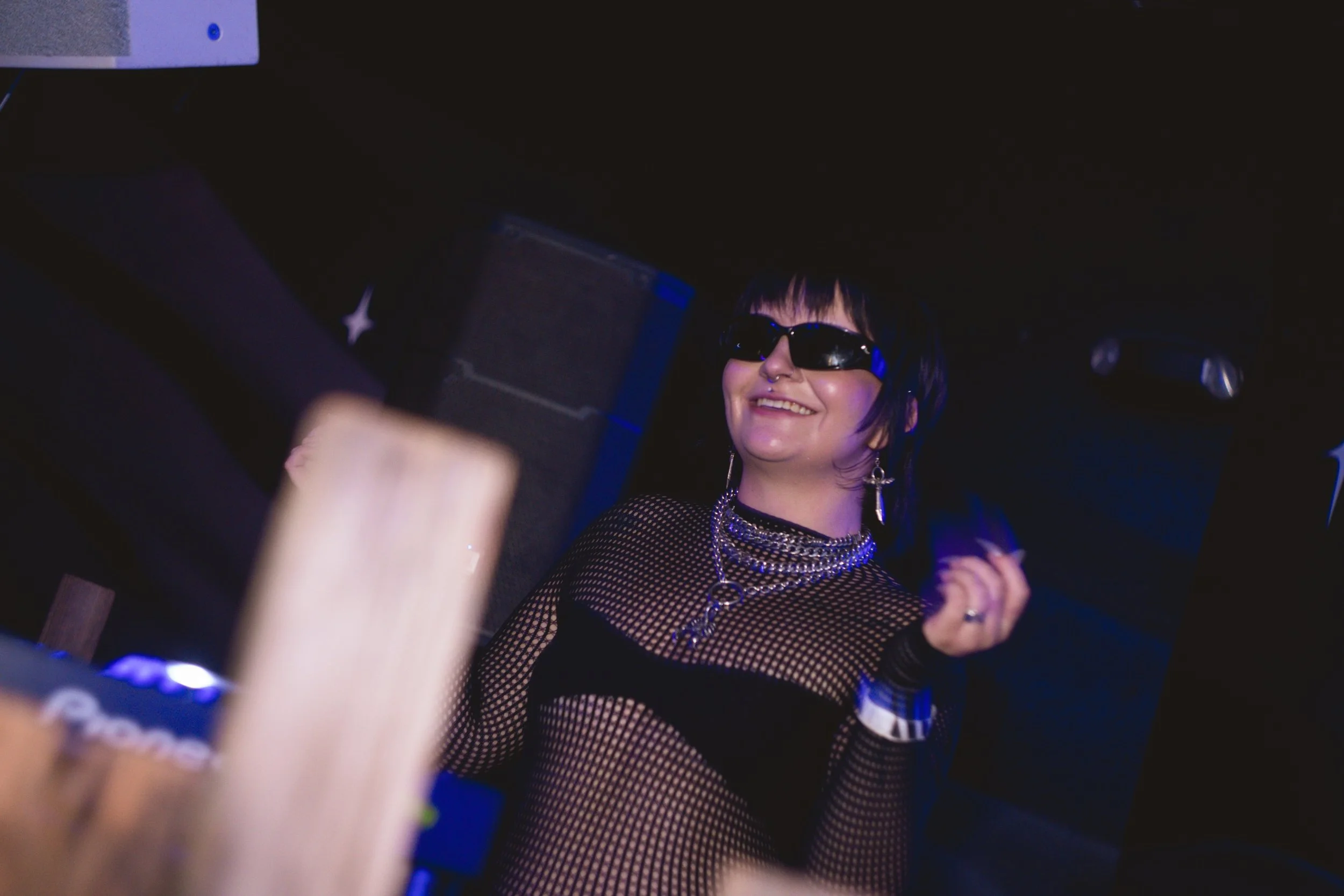 A smiling woman with short black hair and sunglasses, wearing a fishnet top and multiple layered necklaces, in a dark club environment.