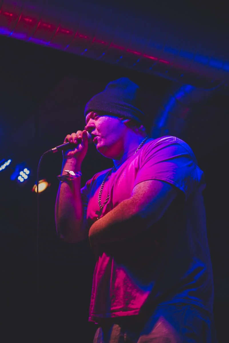 A person wearing a beanie hat and a t-shirt, holding a microphone and performing on stage under colorful lighting.