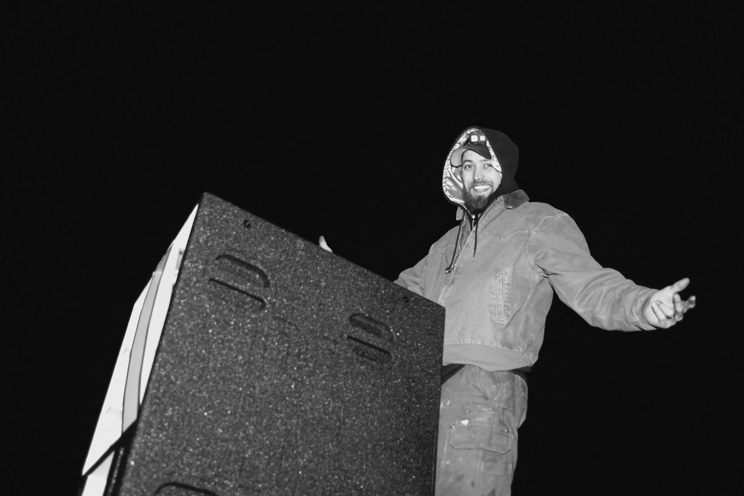 A man smiling and gesturing towards the camera at night, standing behind a speaker or equipment, wearing a jacket and hoodie.