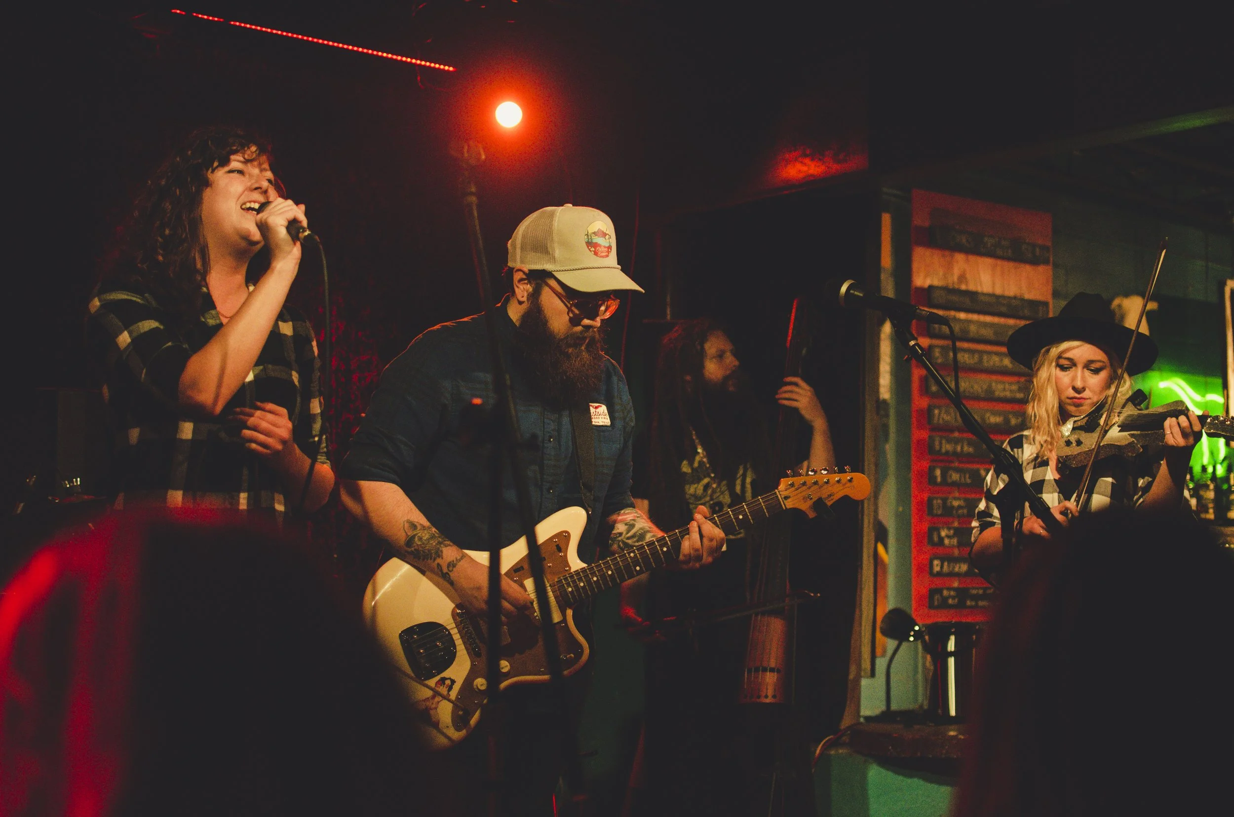 A band performing live on stage in a dimly lit venue, with a female singer with curly hair and a plaid shirt, a male guitarist with a beard and hat, and another female musician playing the violin, all engaged in their performance.