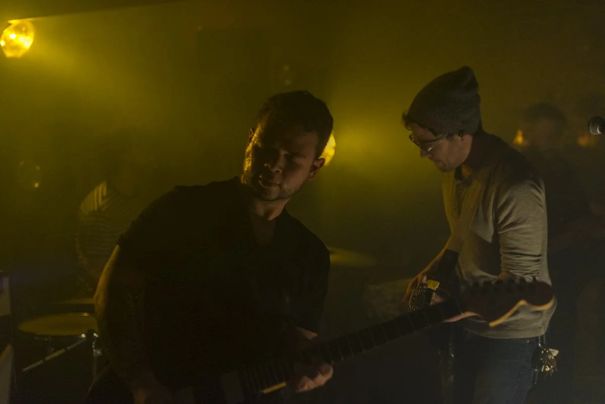Two men playing guitar in a dimly lit setting with yellow lighting, one wearing a beanie and glasses, the other with short hair.