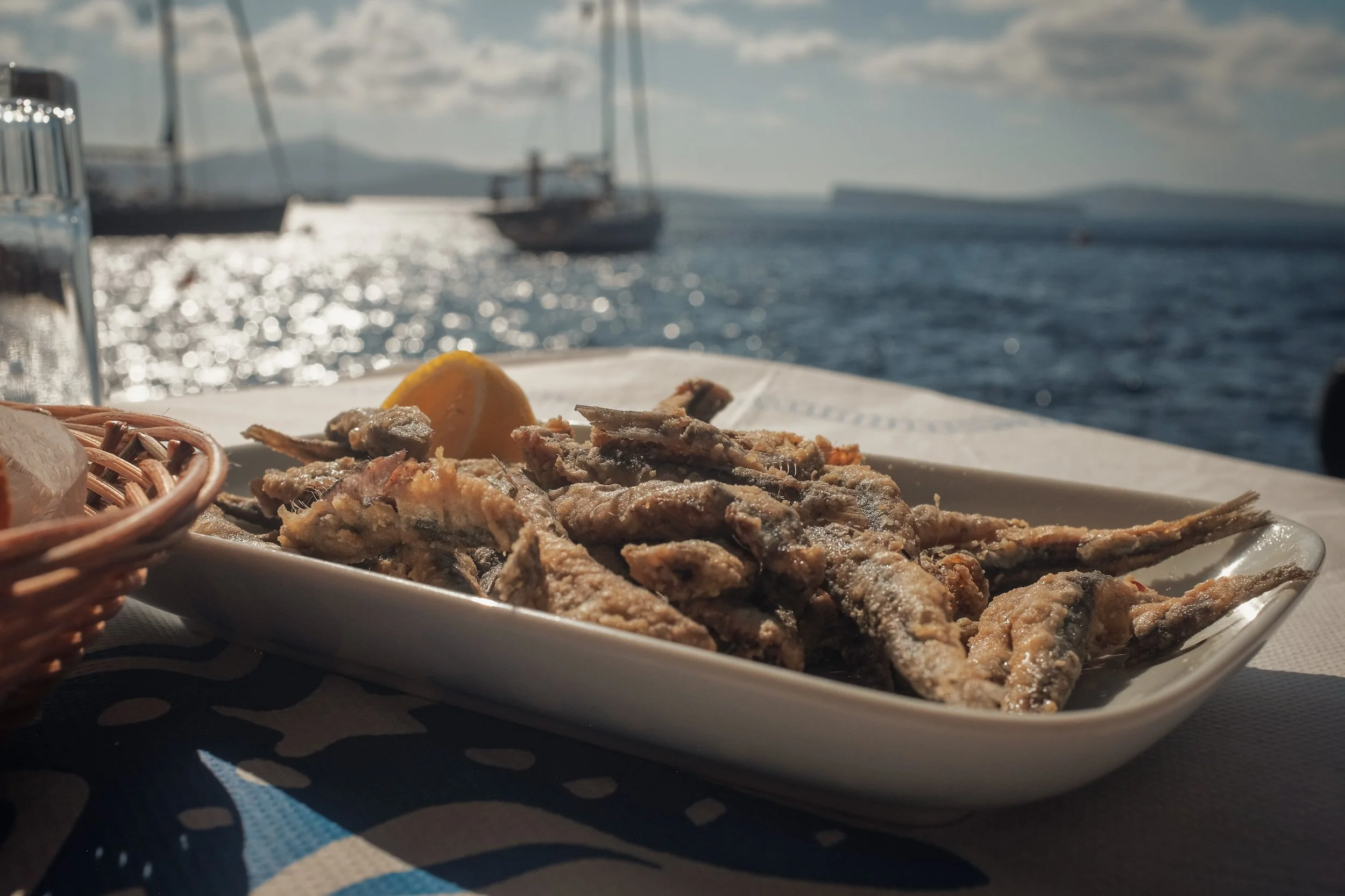 Fried small fish on a white rectangular plate with a lemon wedge, set on a table near a body of water with sailboats and mountains in the background.