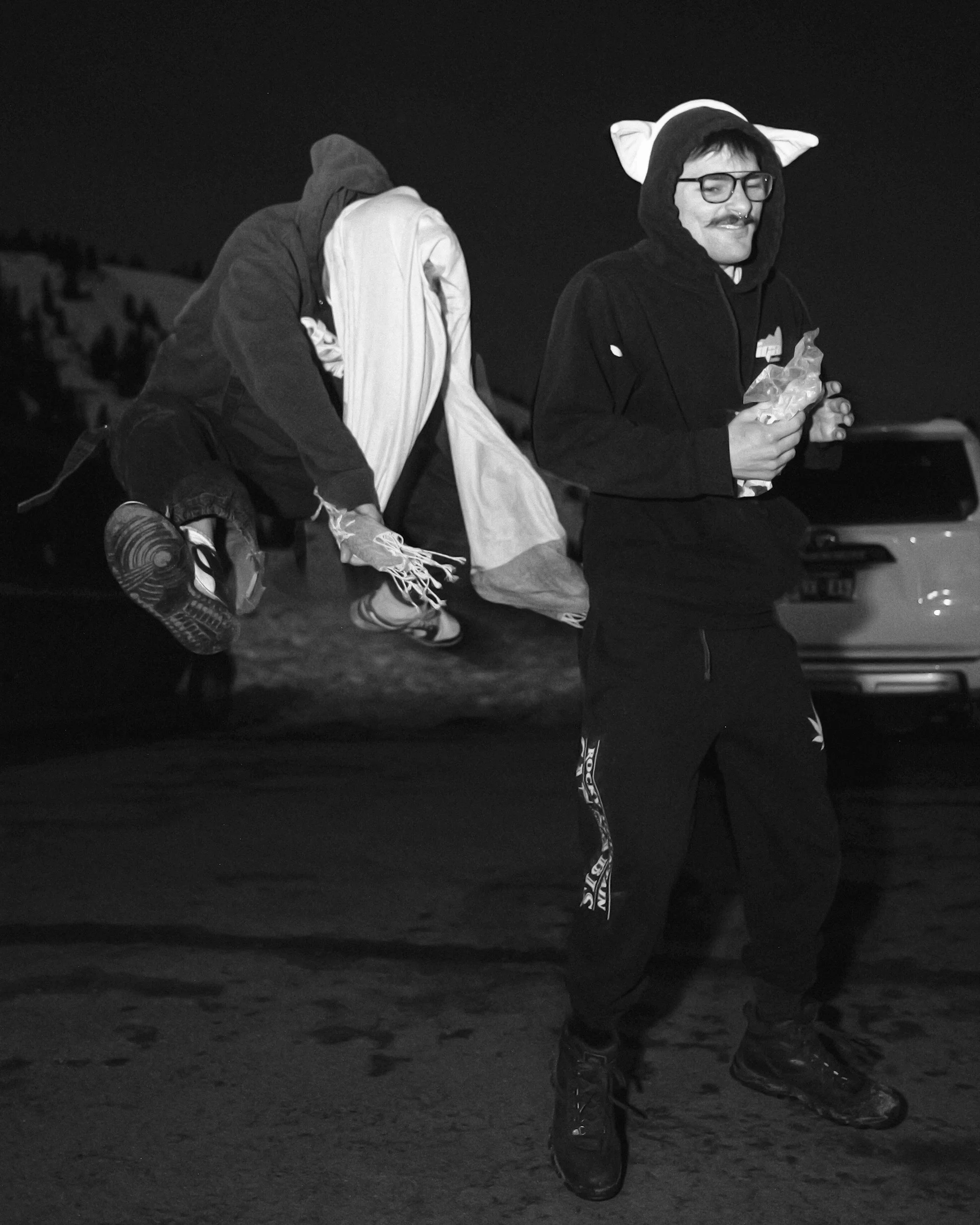 A person wearing glasses and a hoodie with cat ears, holding a snack, stands outdoors at night, with another individual dressed in a hoodie and shirt carrying a large bag.
