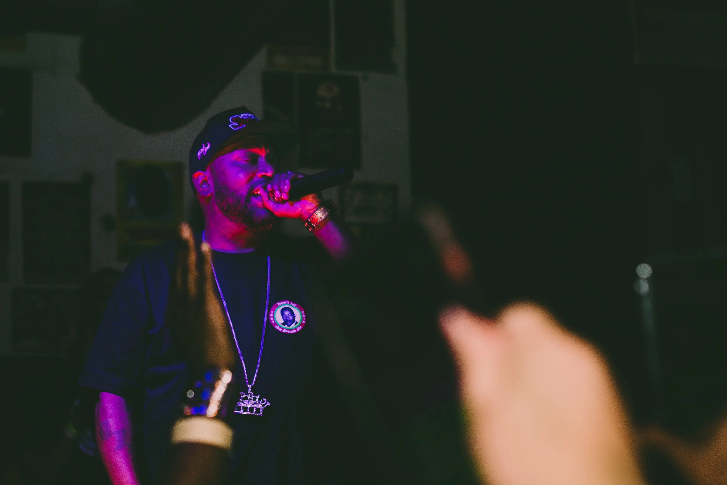 Male rapper performing with microphone in a dimly lit venue, wearing a black cap, black t-shirt with a circular logo, and a chain necklace, with purple and red stage lighting.