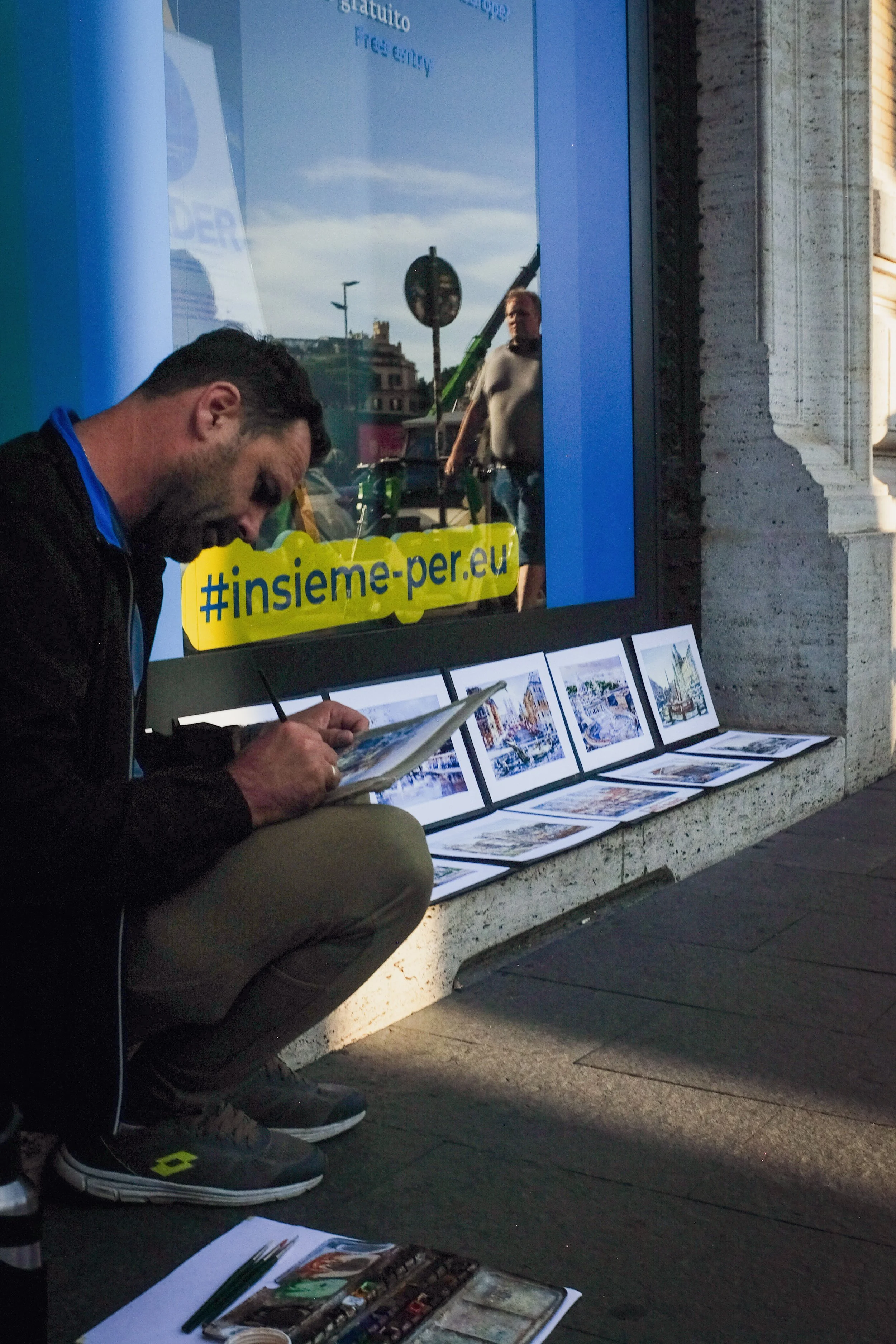 Man crouching on the sidewalk, sketching or painting outdoors, with framed artworks displayed on the ground and leaning against the building behind him, in front of a storefront with a large glass window that reflects a person walking by and a street