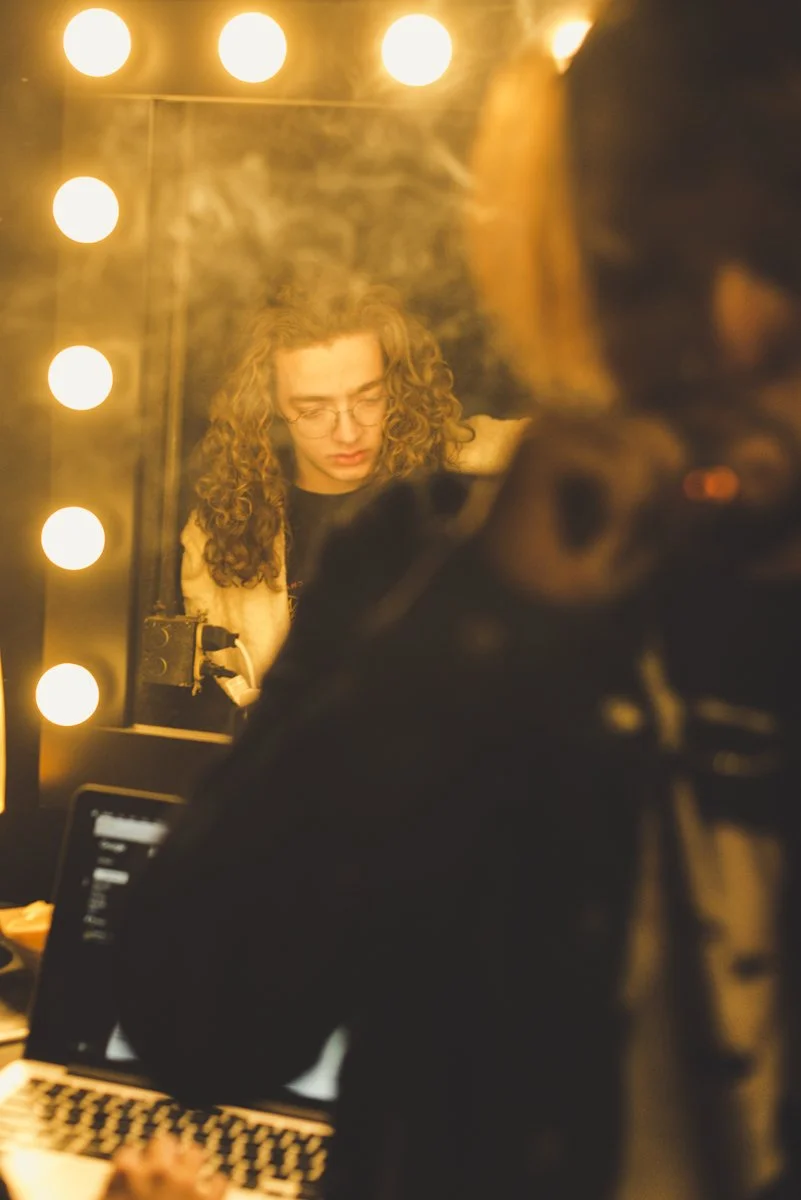 A woman with curly hair and glasses is looking into a mirror with bright lights around it. She appears focused, and there is a person partly visible in the foreground with a laptop and camera.
