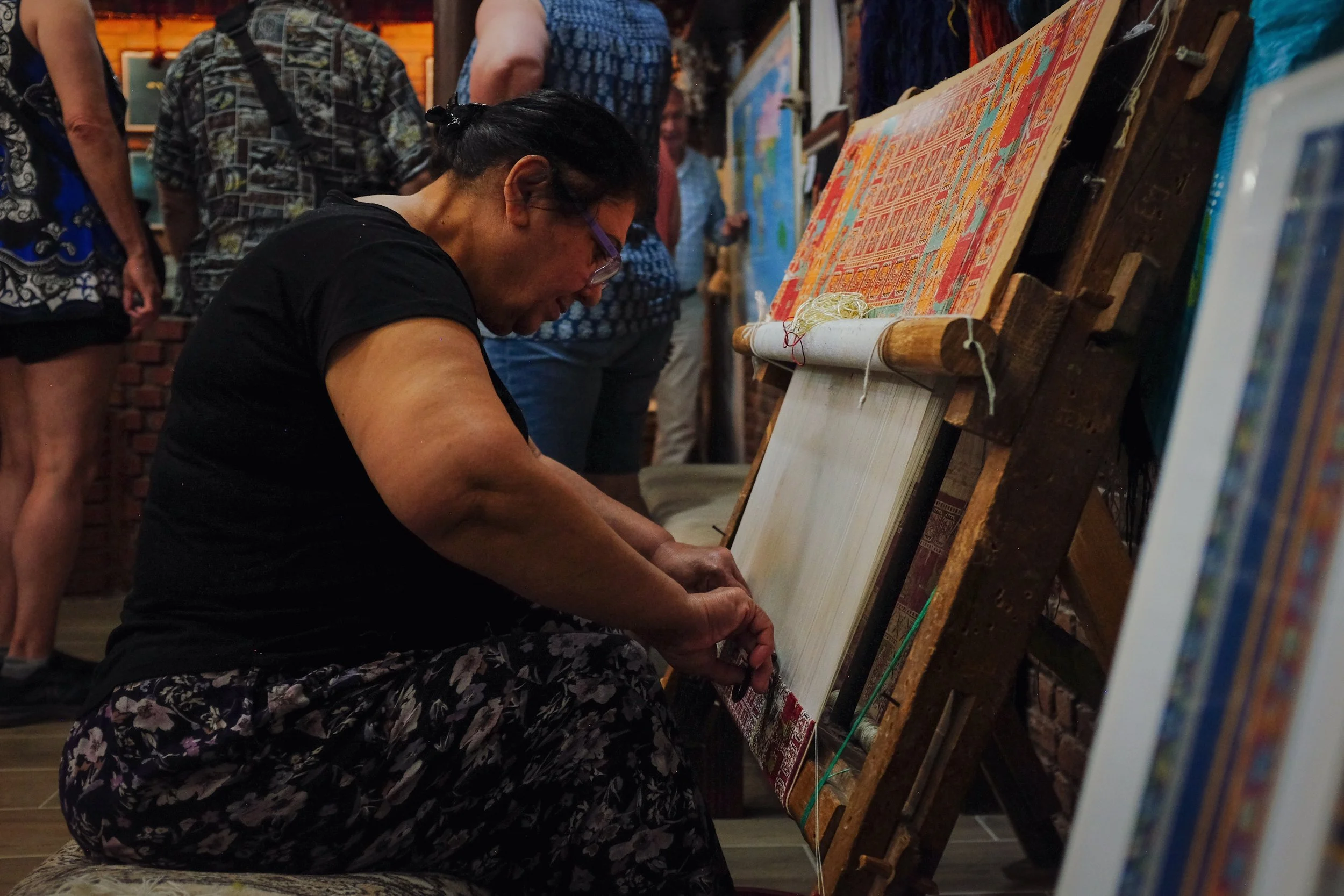 Person weaving on a traditional loom, surrounded by onlookers, in a craft shop or studio.