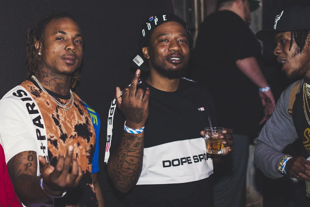 Three young men at a social event, two making hand gestures, one holding a drink, wearing casual streetwear with tattoos and jewelry.