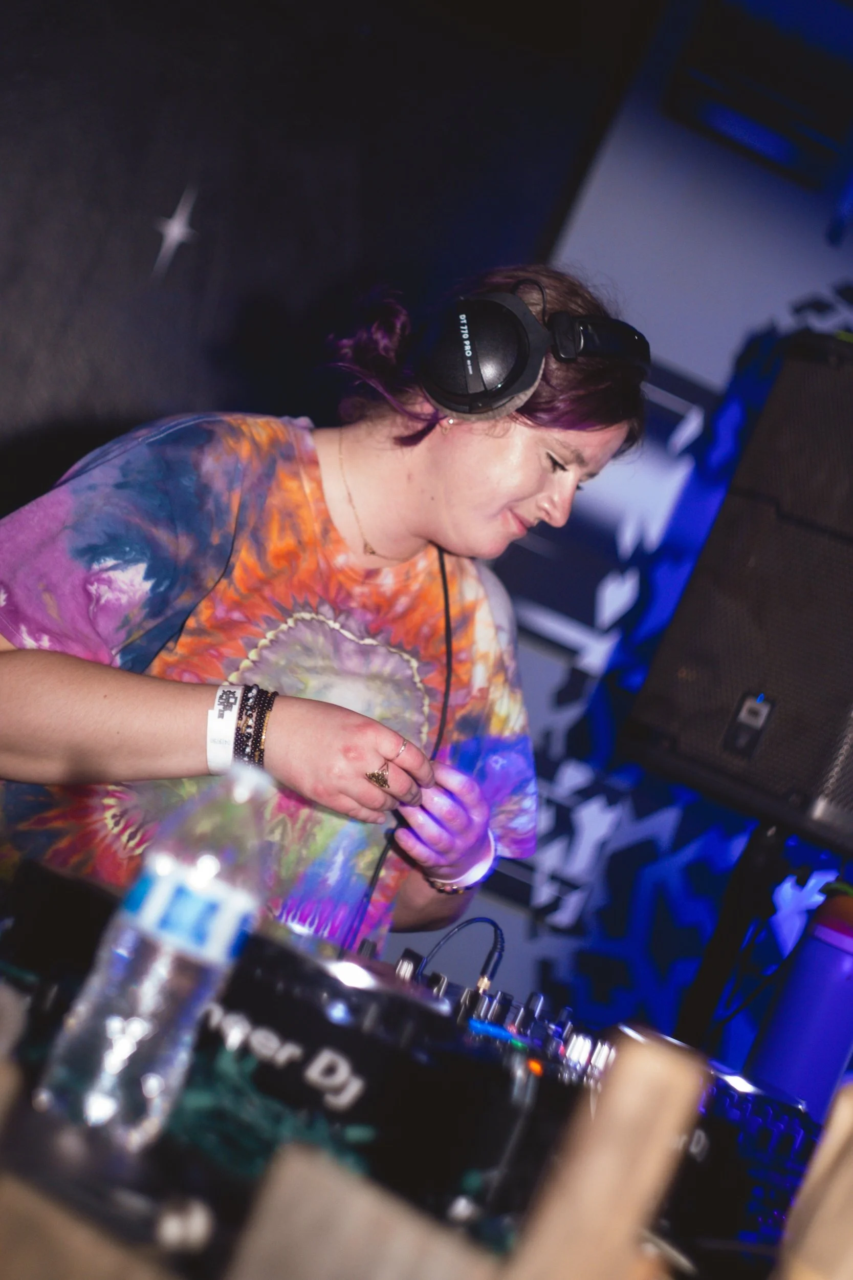 A female DJ with purple hair, wearing headphones and a colorful tie-dye shirt, is standing behind a DJ mixer, smiling while performing at a music event.