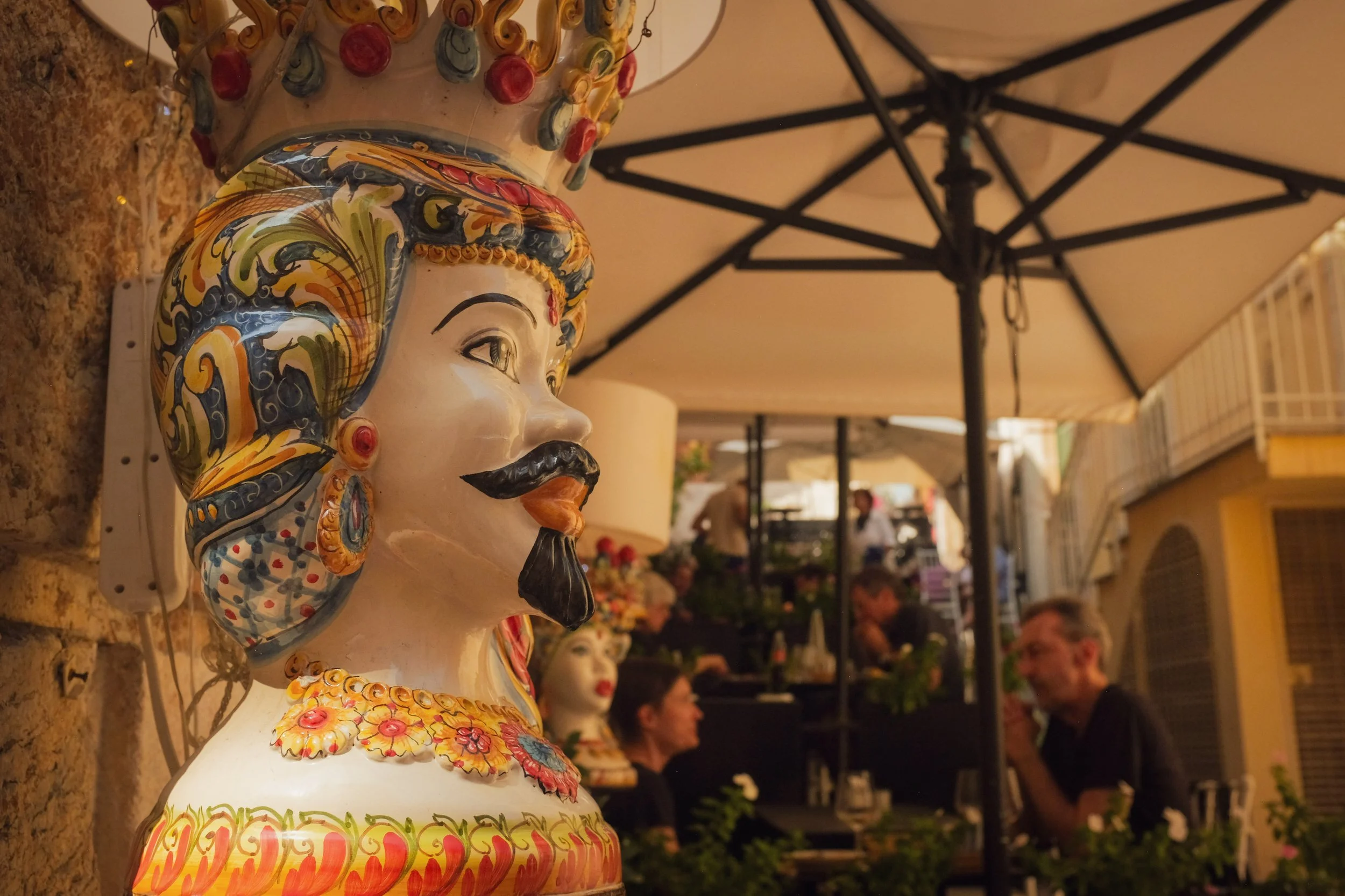 Colorful ceramic bust of a man with a mustache and traditional headdress on a restaurant patio with people dining in the background.