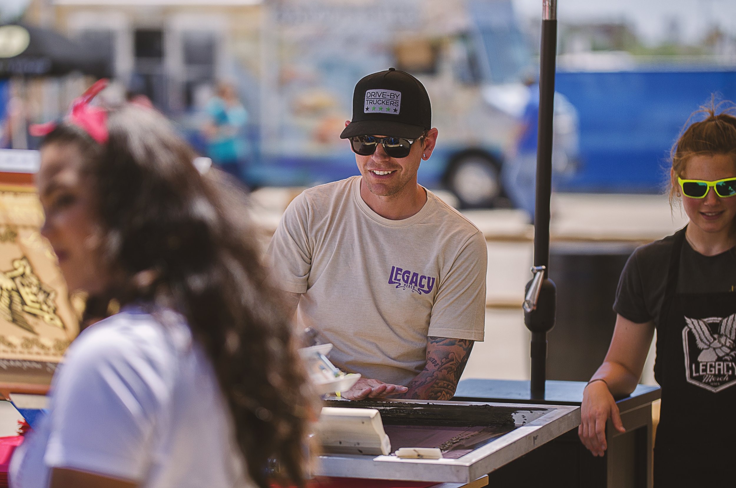 A man wearing sunglasses, a black cap with the words Drive-By Truckers, and a beige T-shirt with the word Legacy, is sitting at a booth and smiling. There are women around him, one in front with dark curly hair and another to his right with blonde ha