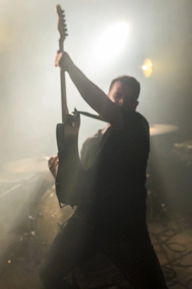A person playing an electric guitar on stage in a smoky, dimly lit environment.