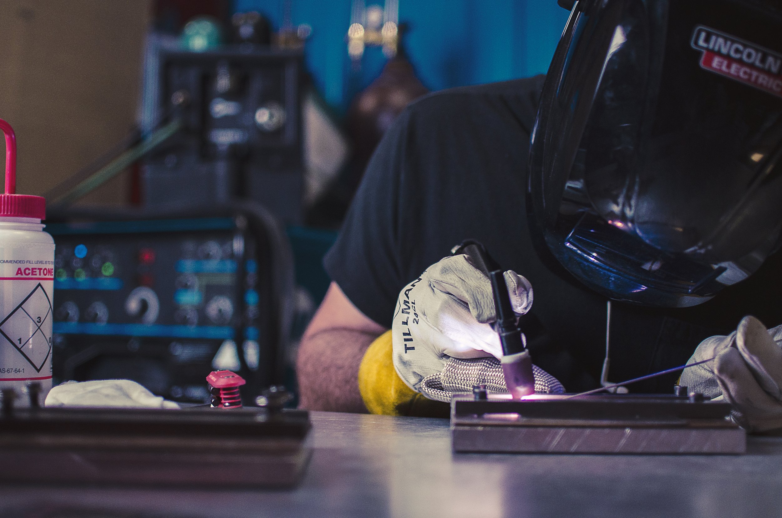A person welding metal on a workbench, wearing a black welding helmet and gloves, with a welding torch in hand, in an industrial workshop setting.