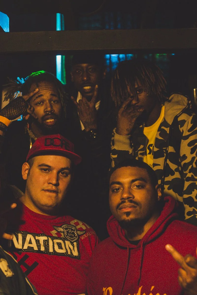 Five young men pose together in a dimly lit environment, some with dreadlocks and casual streetwear, making hand gestures and facial expressions for the camera.