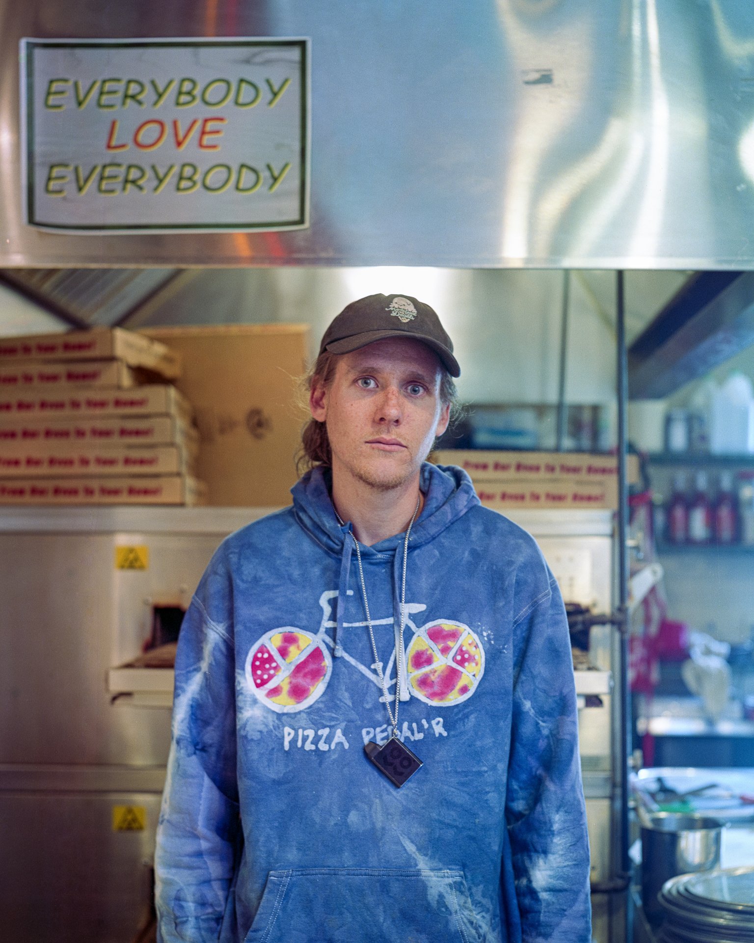 A young man with blonde hair and a black cap standing in front of a pizza shop counter, wearing a blue hoodie with a pizza bicycle graphic and the words 'PIZZA PEDAL'R.' The background shows pizza boxes, a metal kitchen shelf, and various kitchen ite