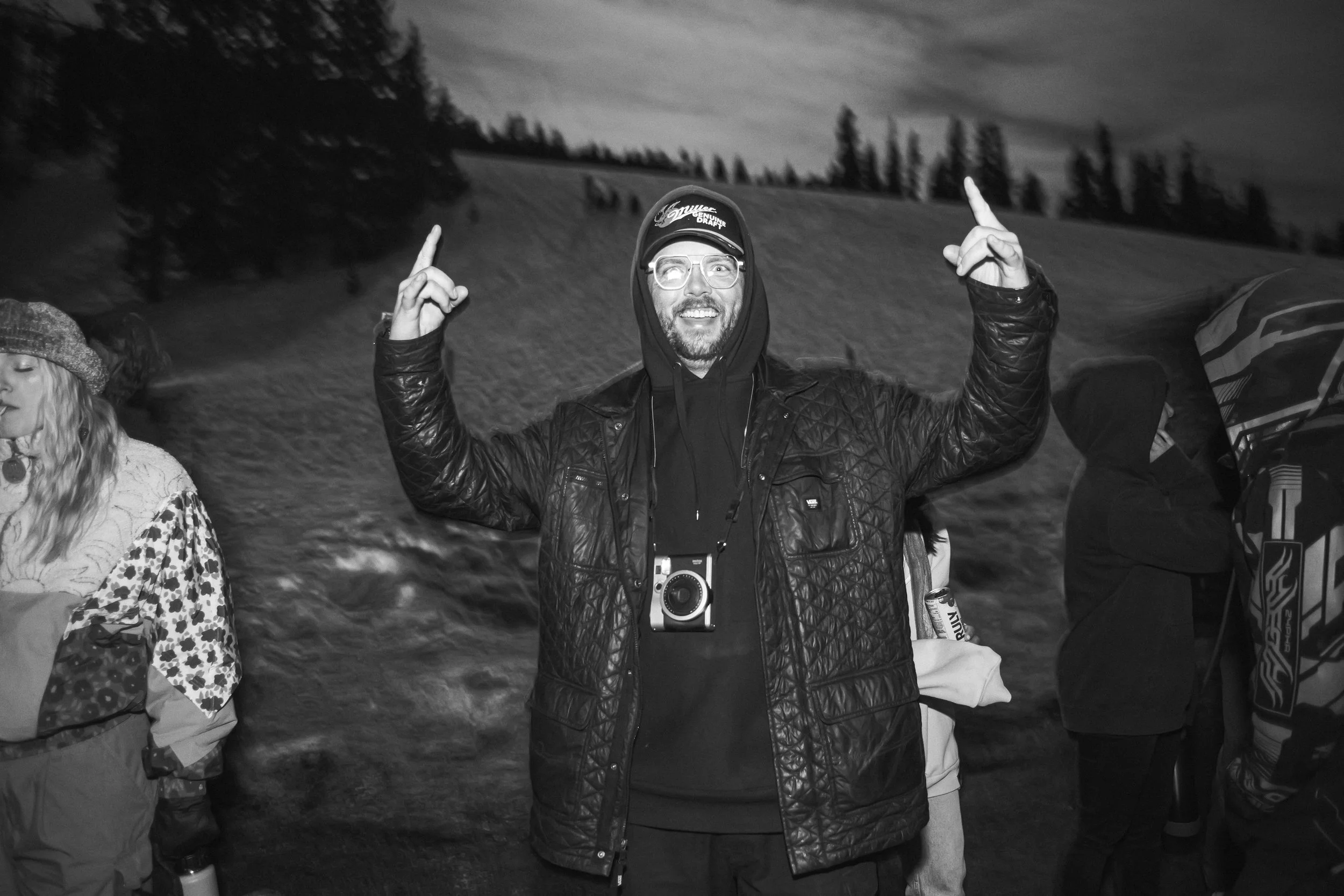 A man smiling with arms raised pointing upward, wearing glasses, a baseball cap, a hoodie, and a quilted jacket, standing outdoors at night with snow-covered ground and trees in the background.