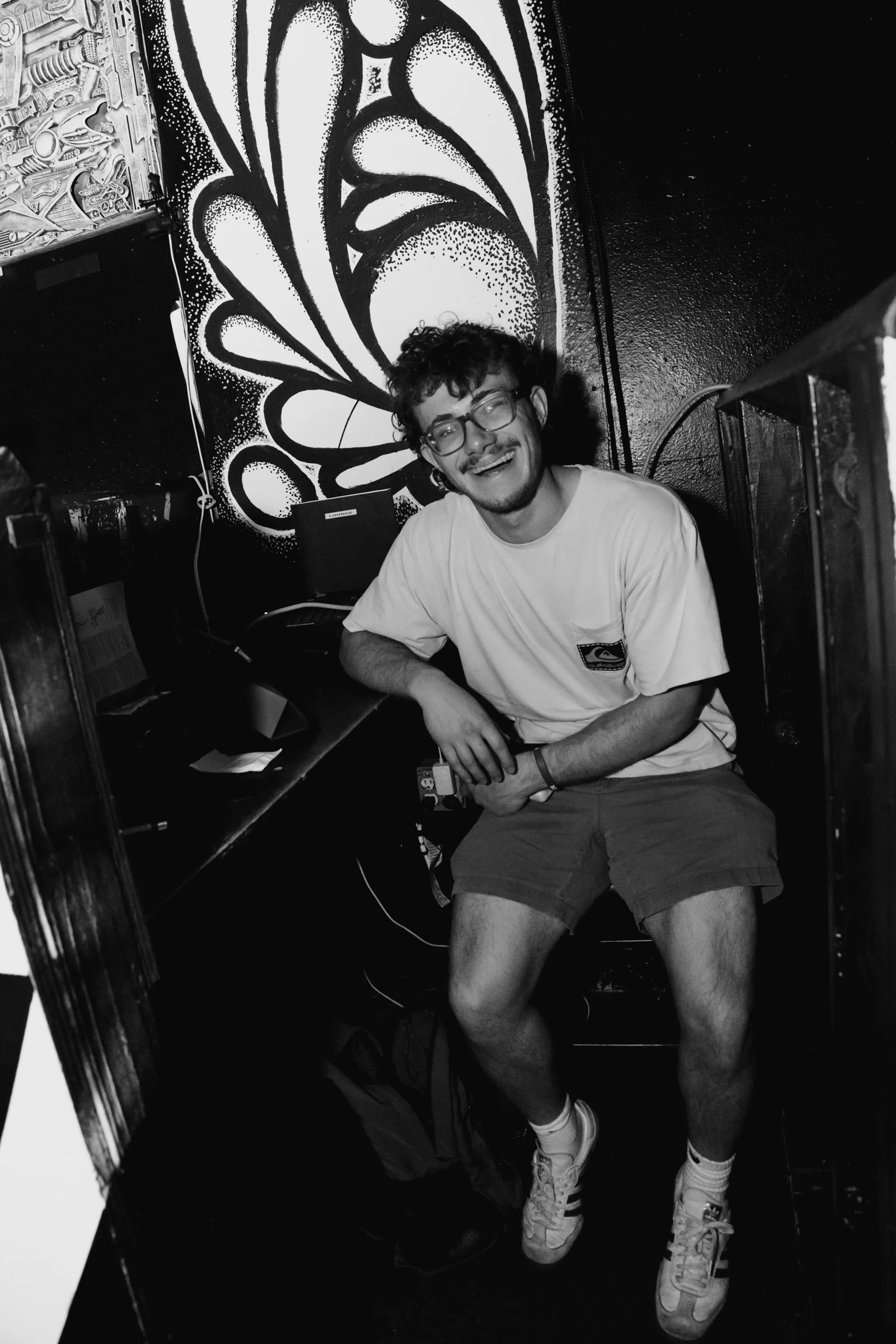 A young man with curly hair and glasses sitting and smiling in a room with a large butterfly wing mural painted on the wall behind him. He is wearing a t-shirt, shorts, and sneakers.