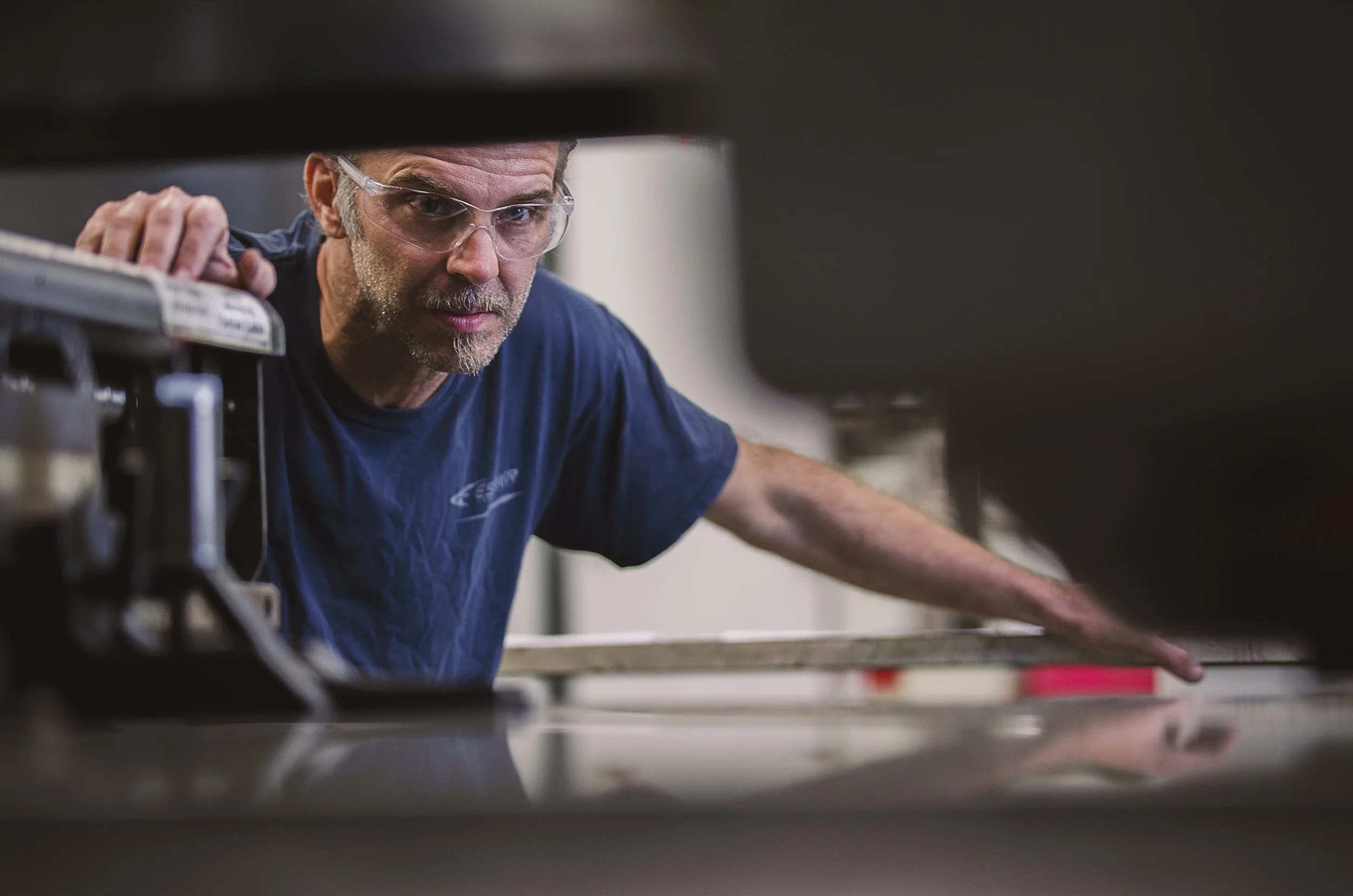 A man wearing safety glasses and a navy blue t-shirt leaning over a workbench in a workshop or factory.