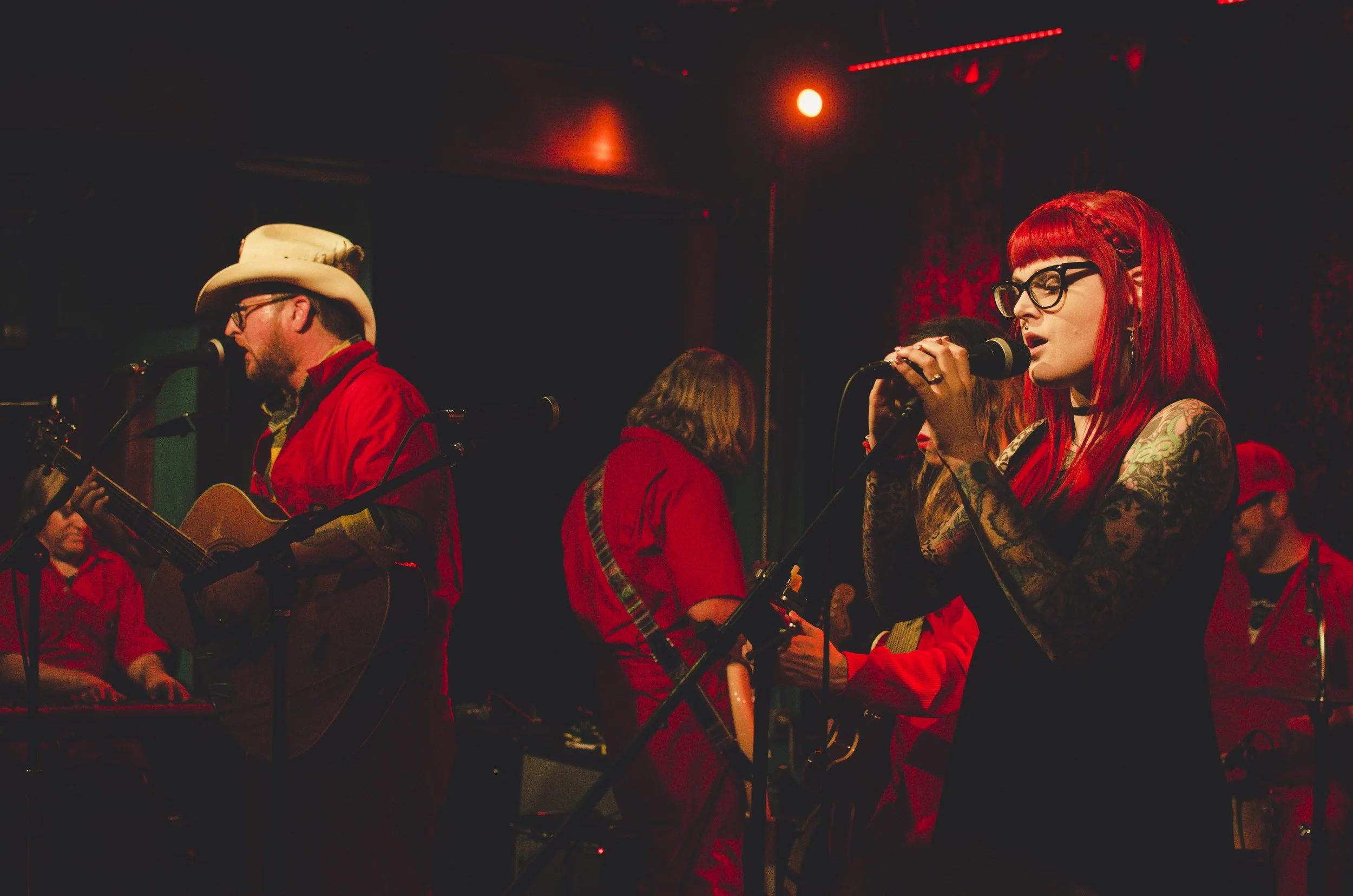 A band performing on stage with a woman with red hair, tattoos, and glasses singing into a microphone, accompanied by male musicians playing guitar and keyboard, all dressed in red attire.