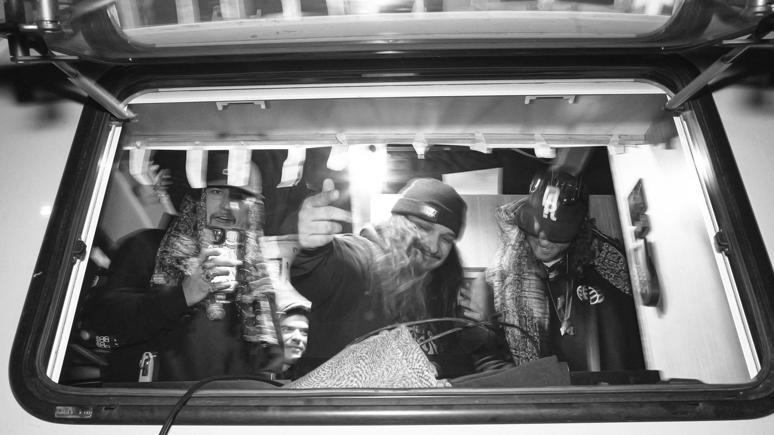 Four people are taking a selfie in a small enclosed space, possibly a bus or van, visible through a window. One person is holding a phone, and another is wearing headphones and a cap. They are smiling and appear to be having fun.