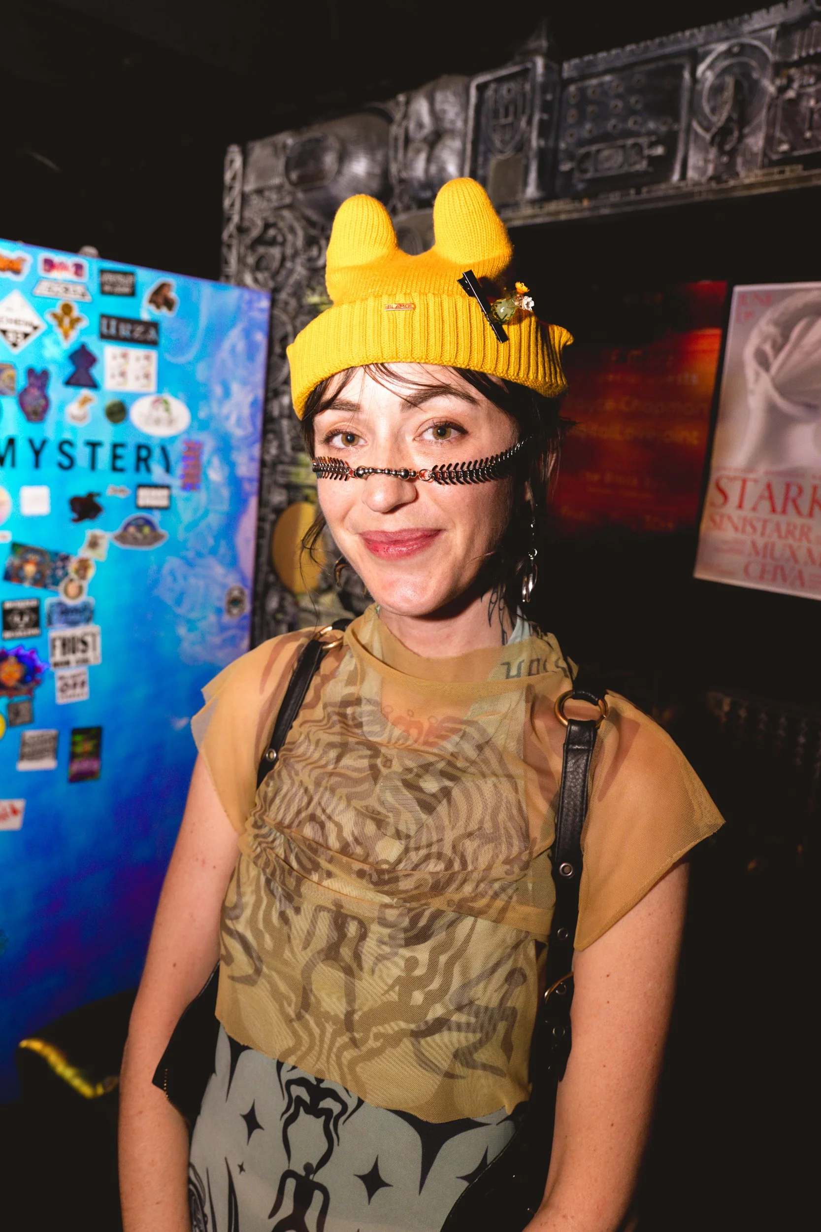 A woman with short dark hair wearing a yellow hat with ears, a chain eye glasses accessory, a sheer yellow top with black patterns, and earrings. She stands in front of a colorful wall covered with stickers and a dark background with posters.